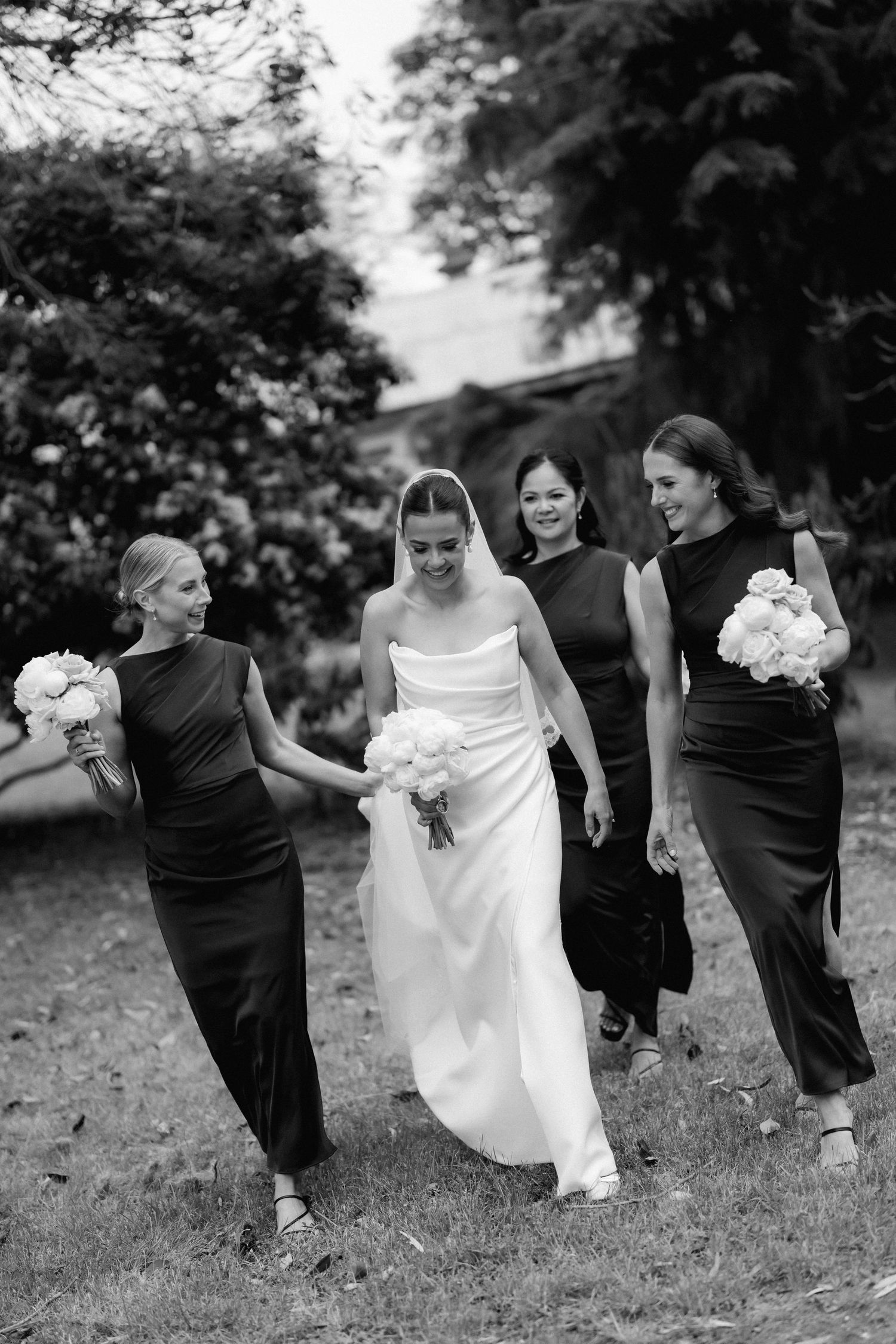 Wedding party members in black and white dresses playfully kicking their legs up on grassy outdoor setting.