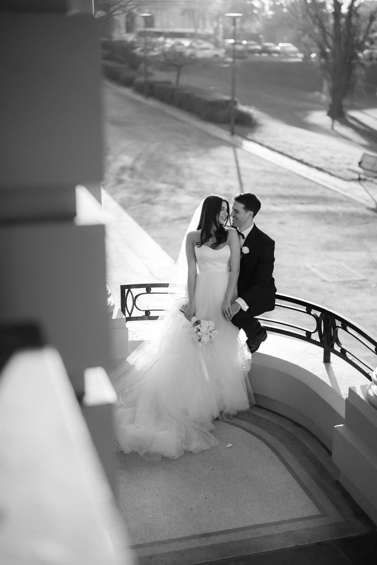 Black and white photo of a bride and groom sharing a romantic moment on an elegant curved balcony staircase.