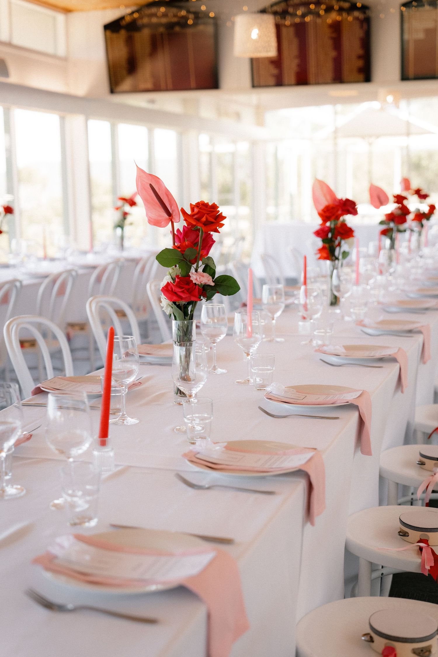 Elegant wedding reception table with red roses, pink napkins, crystal glasses, and red taper candles in a bright white venue.