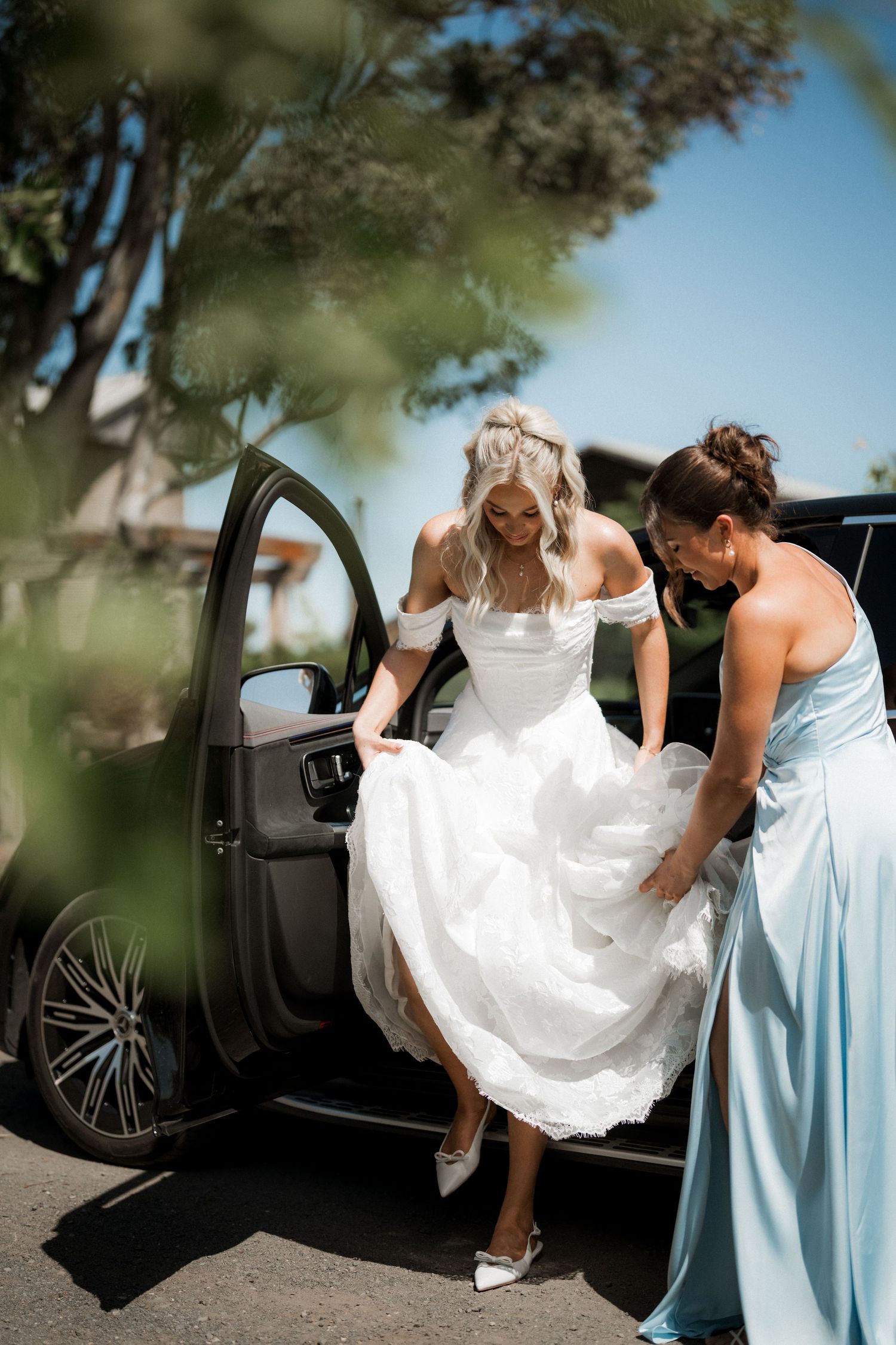 Bridesmaid in light blue dress helping bride in white lace gown exit black car on sunny day.