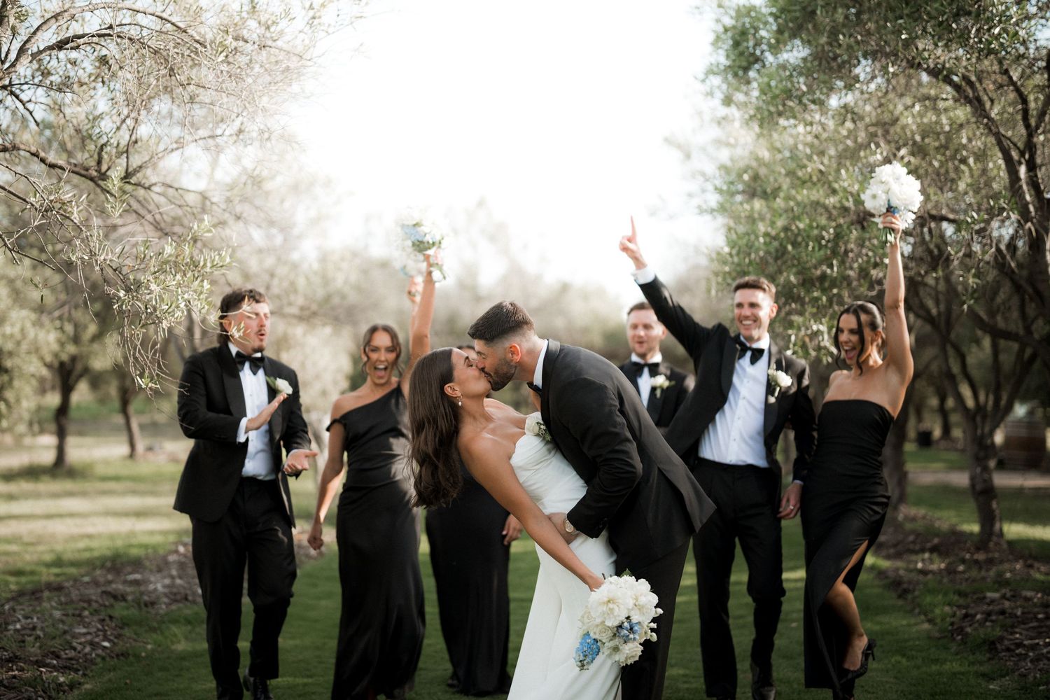 Wedding party celebrates with energetic poses and cheers in an outdoor garden setting.