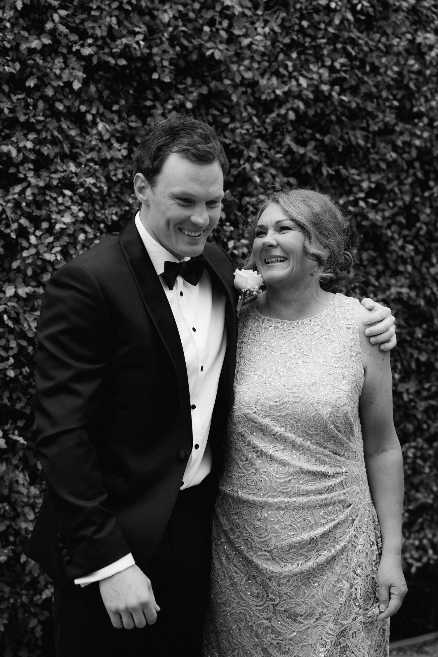 A formal couple sharing a joyful moment against a green hedge backdrop.