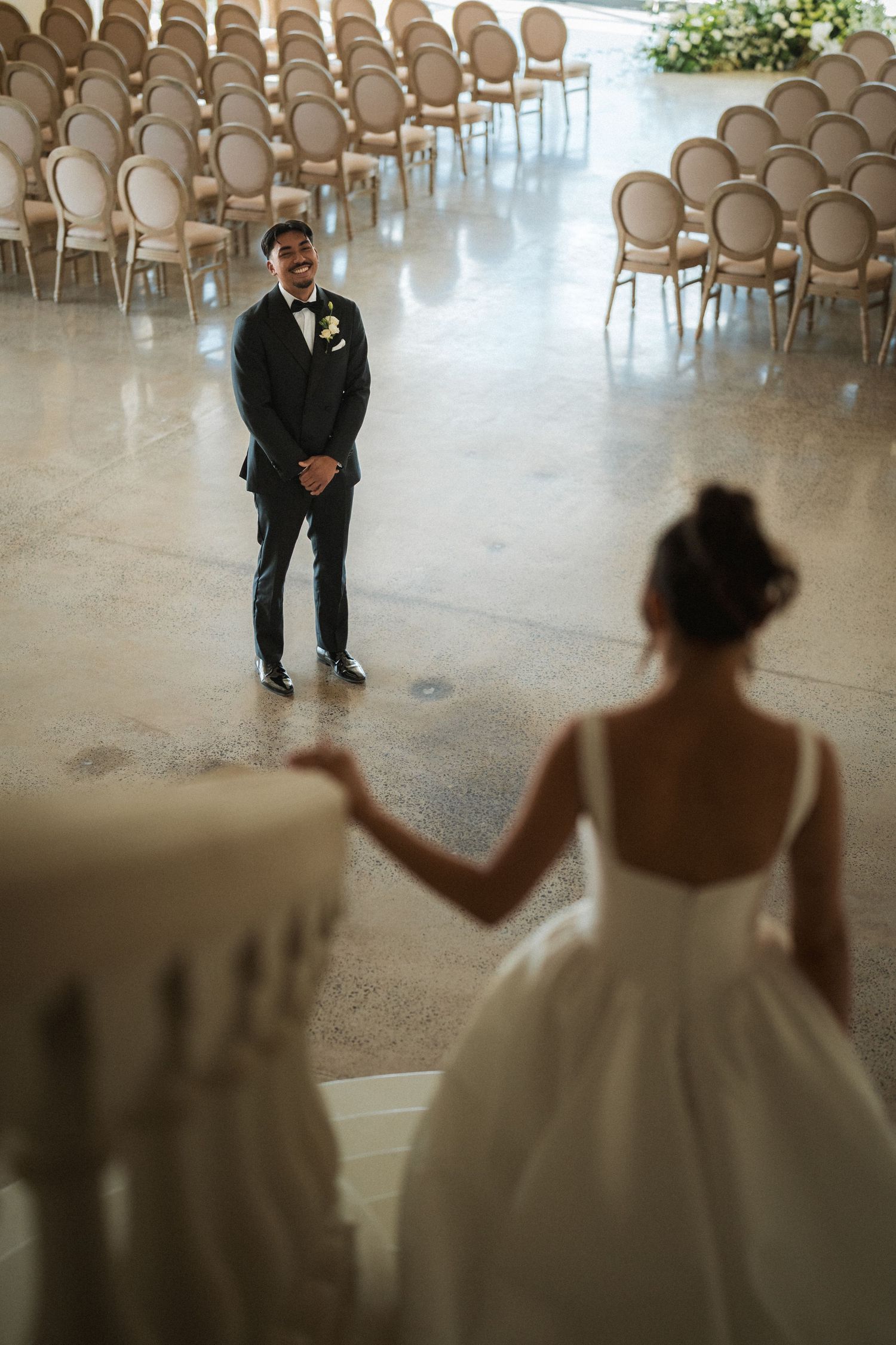 A groom in a black tuxedo smiles in an elegant venue as a bride in a white gown reaches toward him from a staircase.