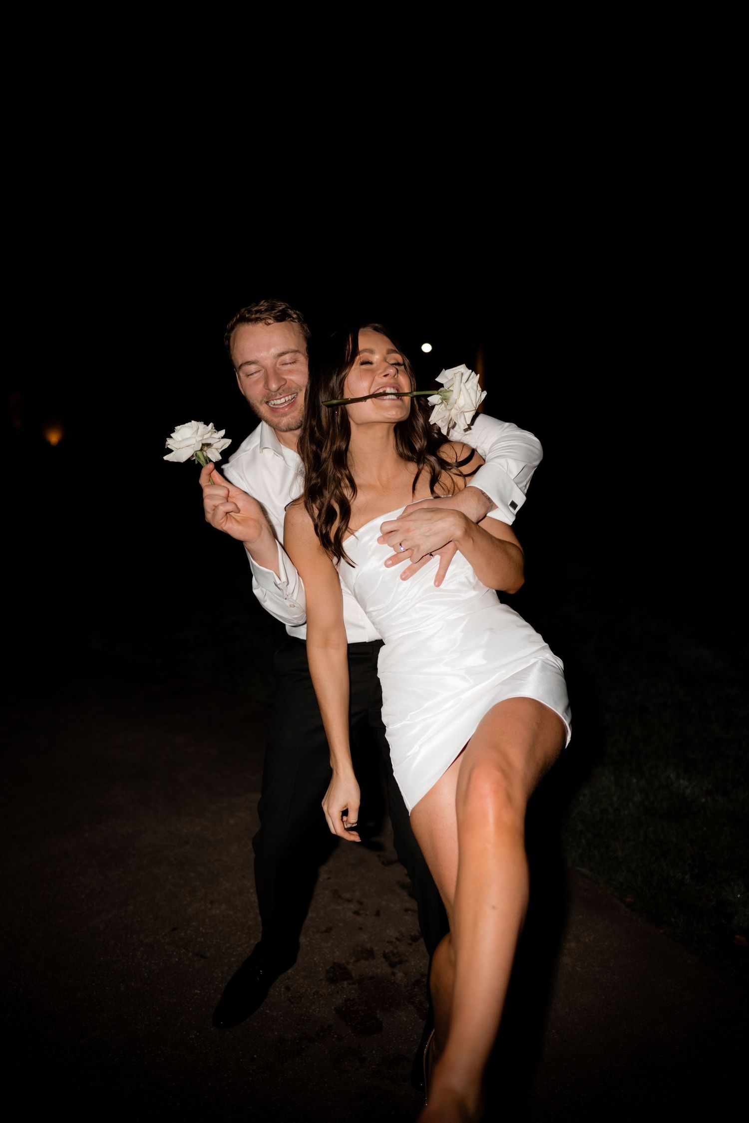A joyful couple in wedding attire share a fun dance moment with white flowers at night.