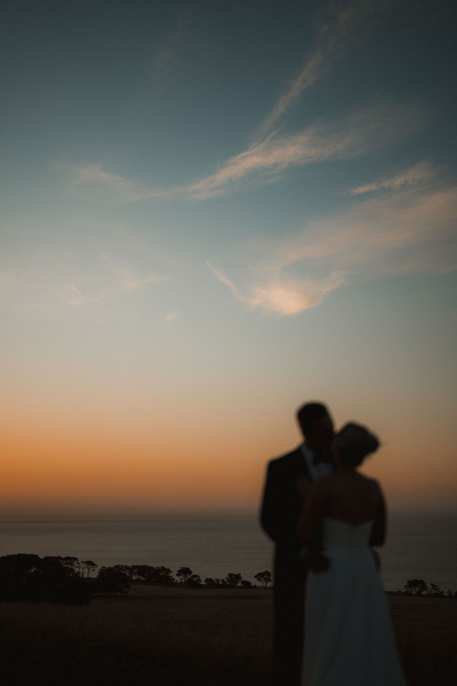 Silhouetted couple embrace against dramatic sunset sky overlooking coastal view.