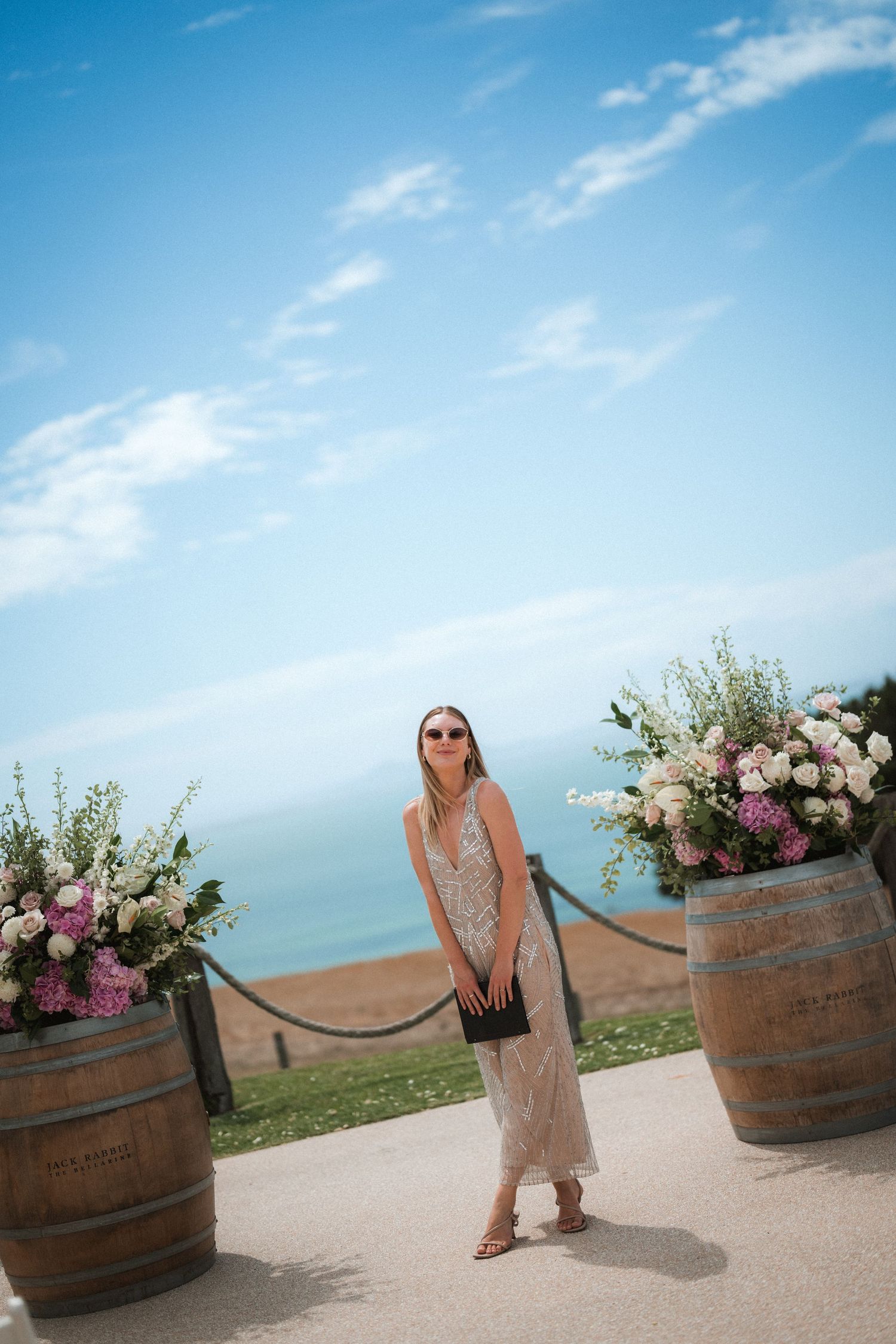 A guest poses between wine barrel planters at an oceanside wedding venue under blue skies.