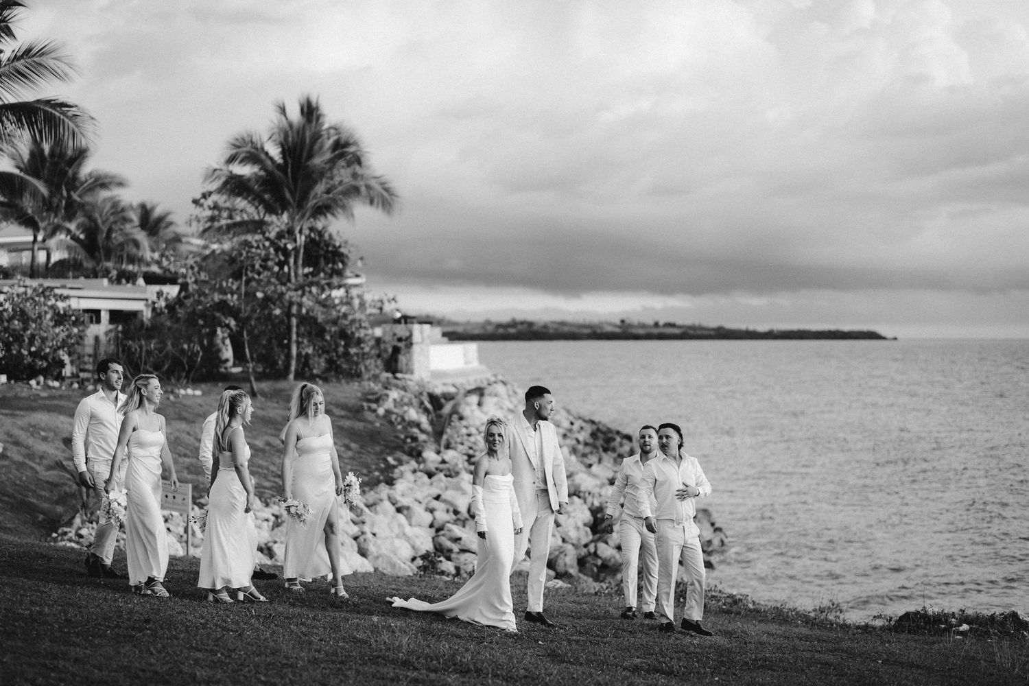 Wedding party in white attire walking along coastal path with palm trees and ocean view.