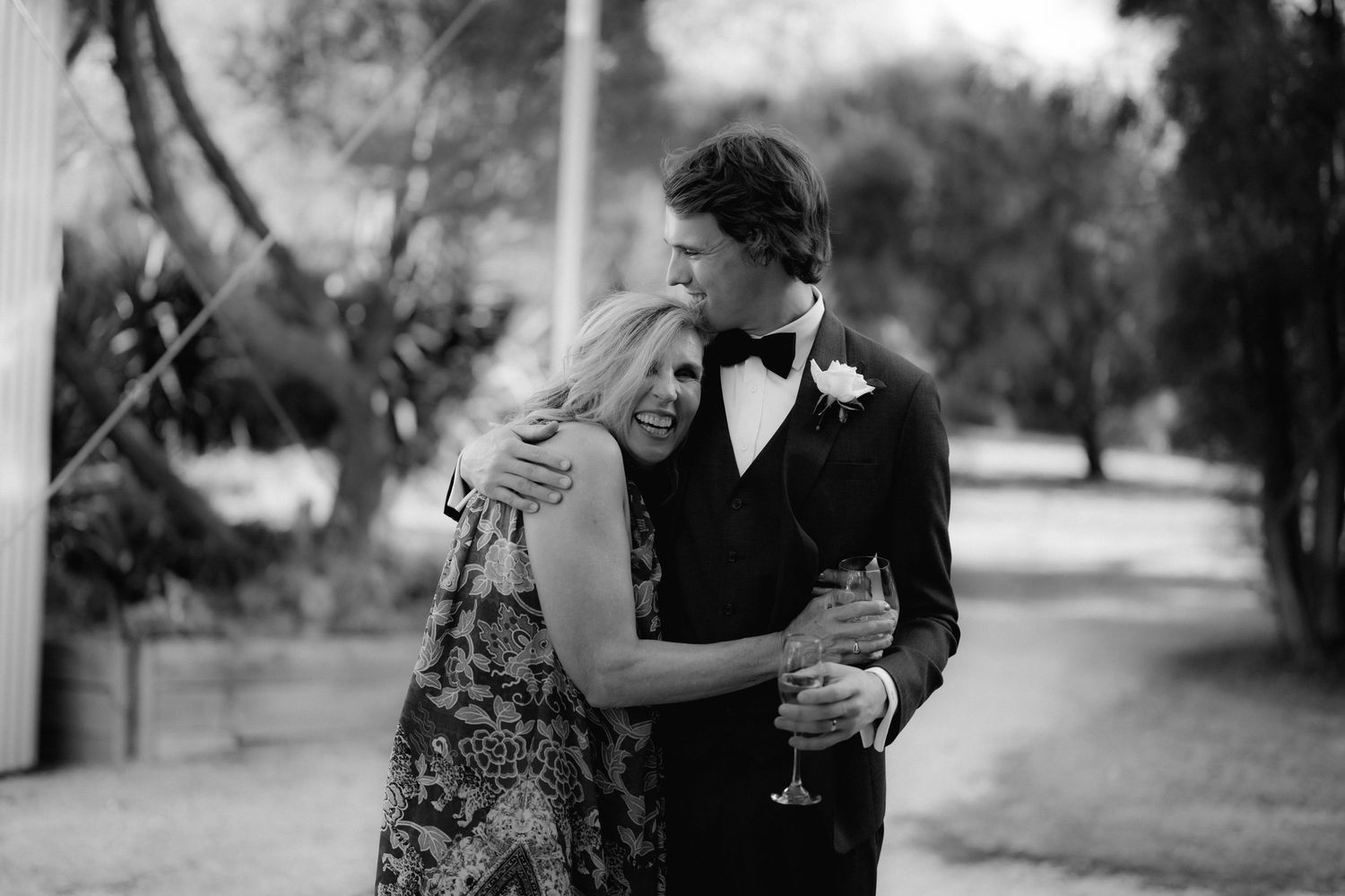 A black and white candid photo of a couple embracing at an outdoor event with wine glasses in hand.