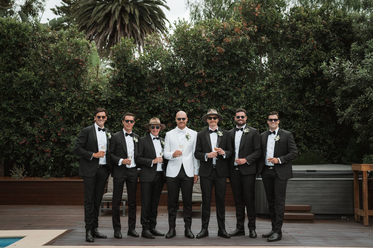 A groom in white tuxedo jacket stands with six groomsmen in black tuxedos against a garden backdrop.