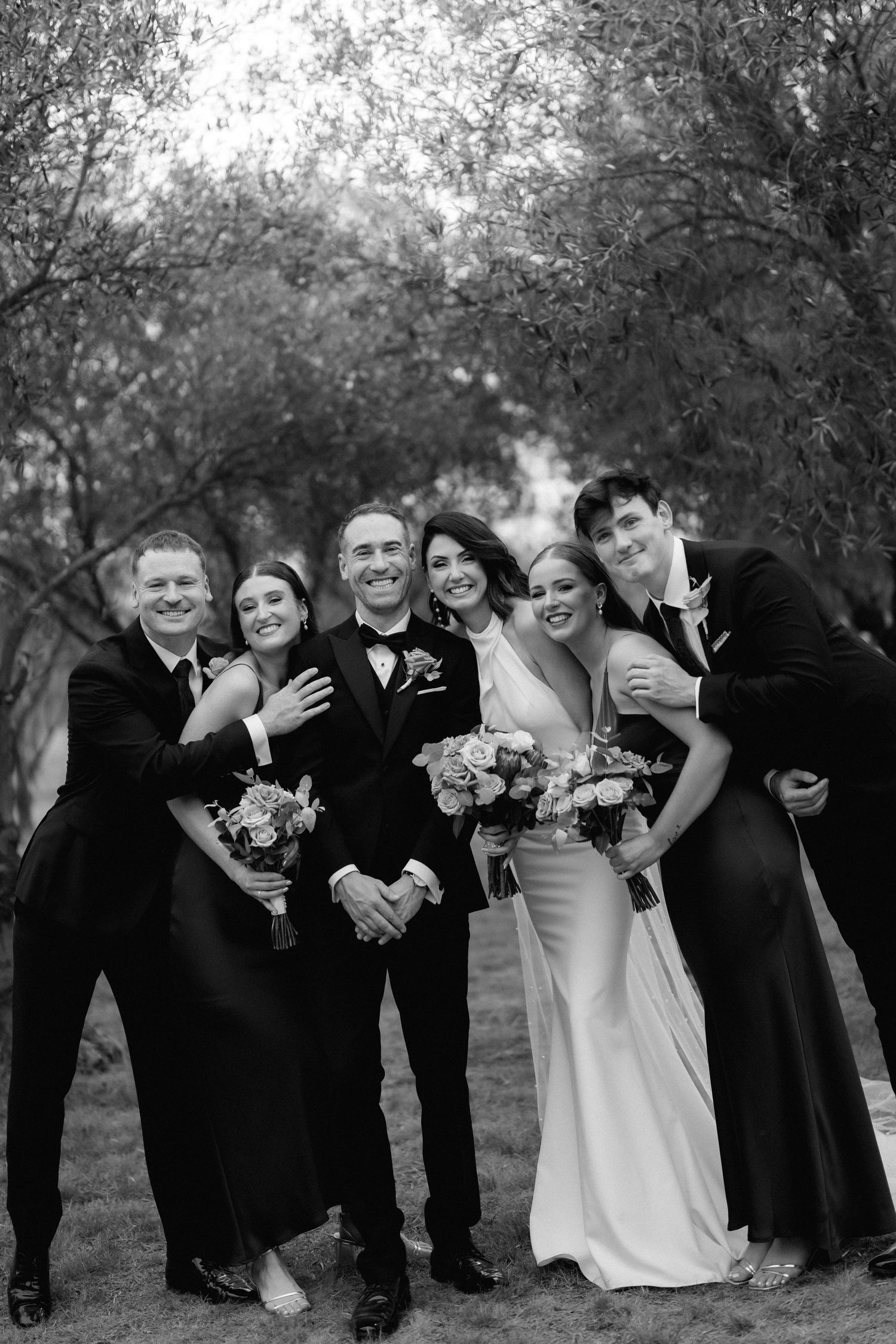 Wedding party in formal black attire poses together outdoors in a candid joyful moment among trees.