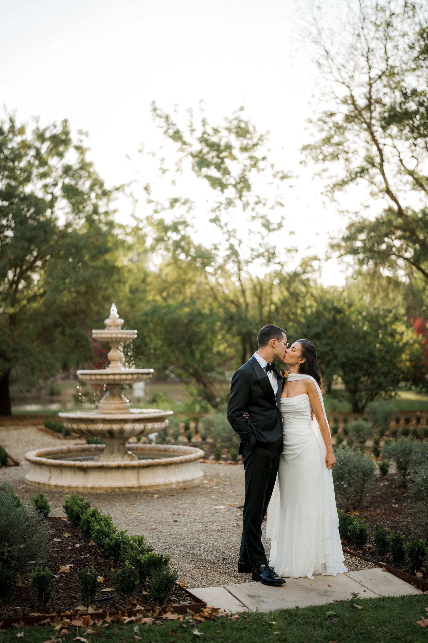 Bride and groom share a romantic kiss near an elegant stone fountain surrounded by lush garden greenery at sunset.