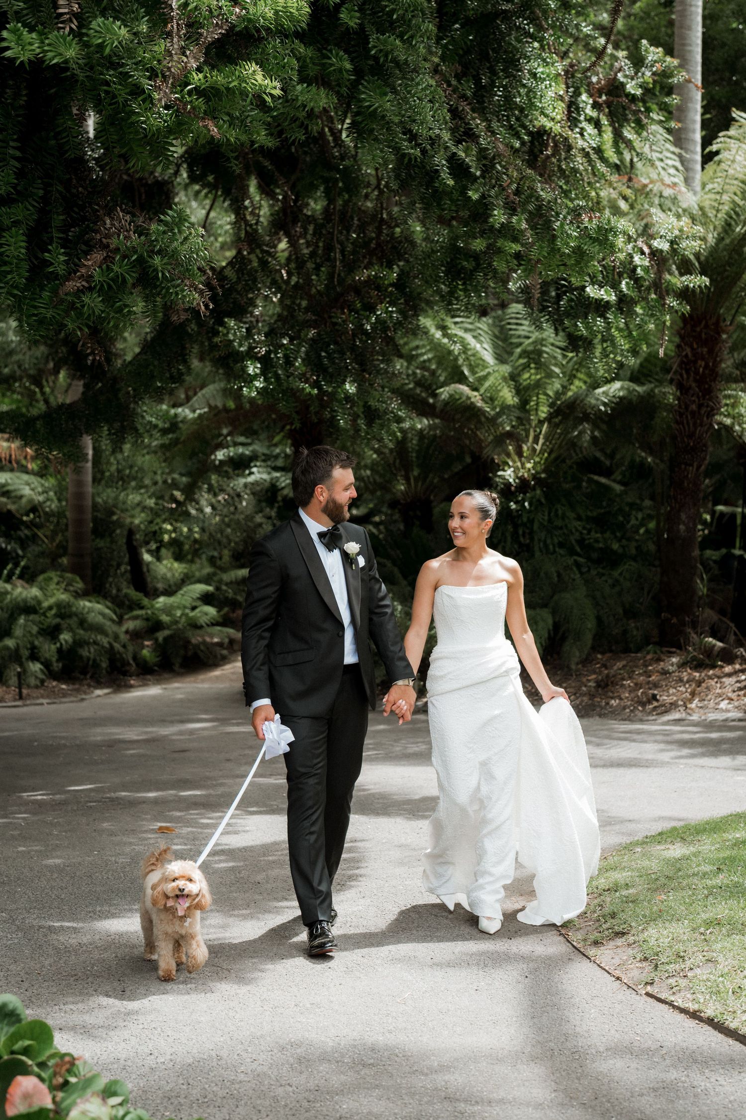 Bride and groom walking hand in hand with their dog on a lush garden path.
