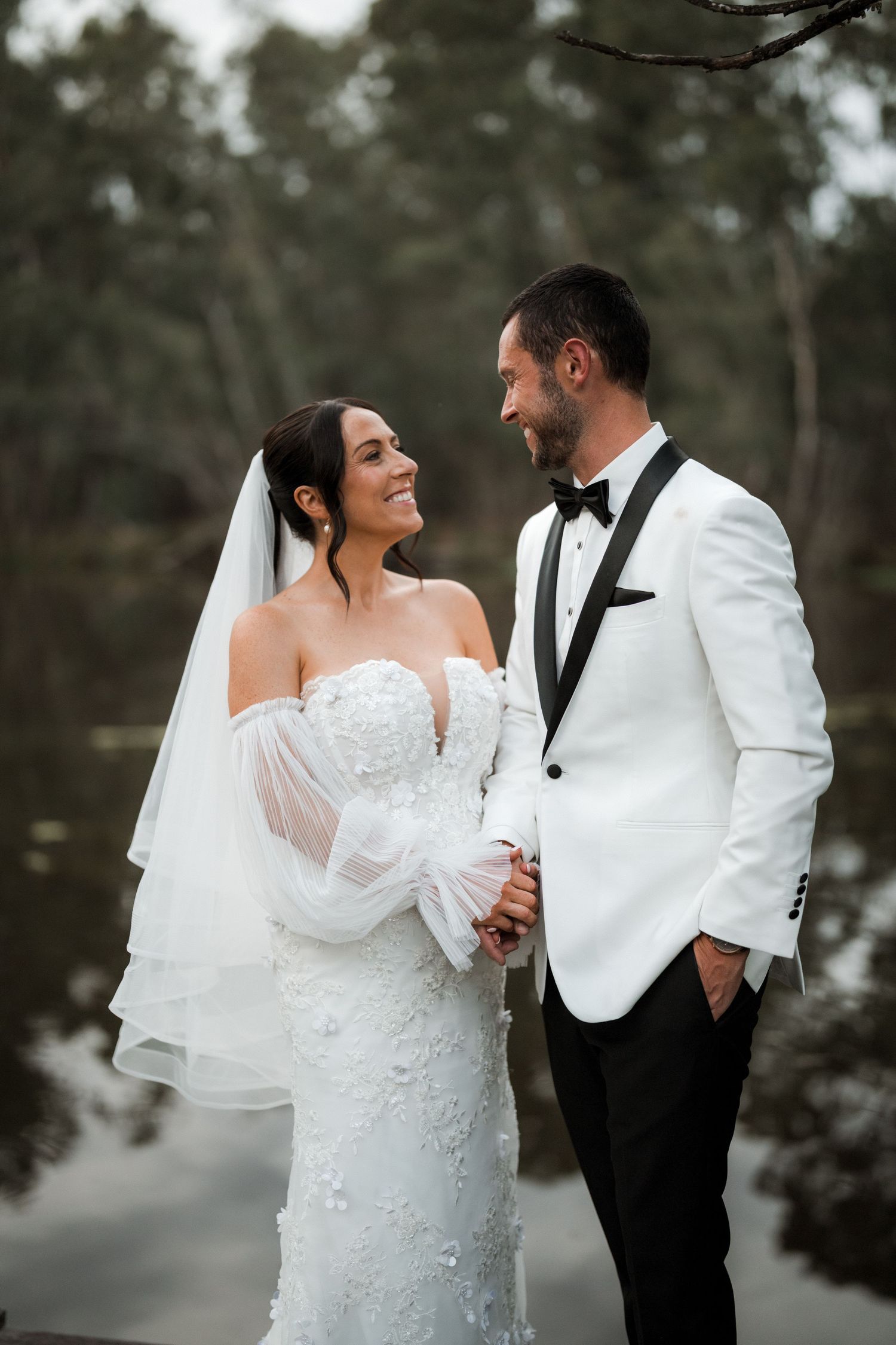 A romantic wedding portrait of a couple in white attire standing against a misty forest backdrop.