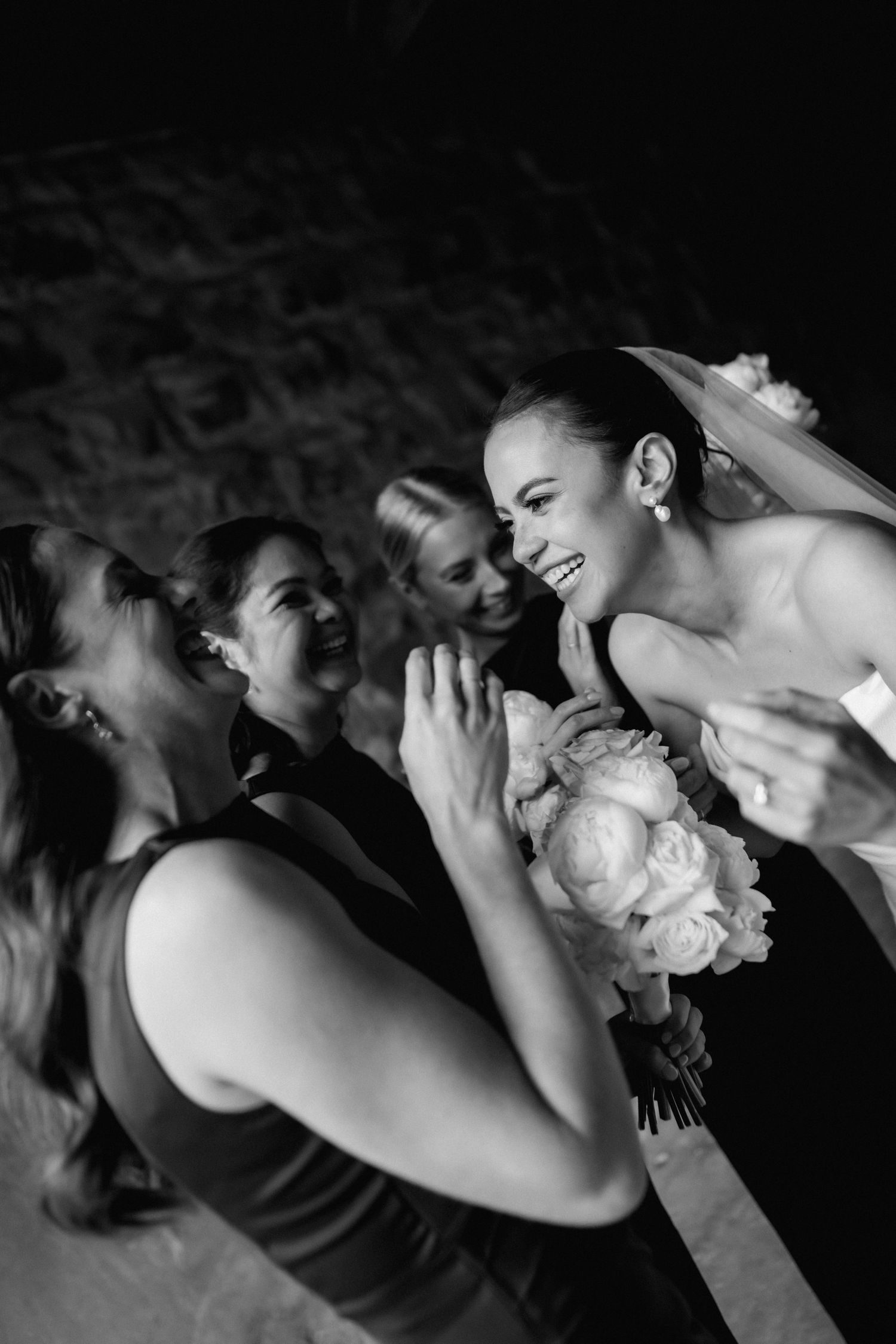 A candid black and white photo captures joyful laughter between a bride and her wedding party.