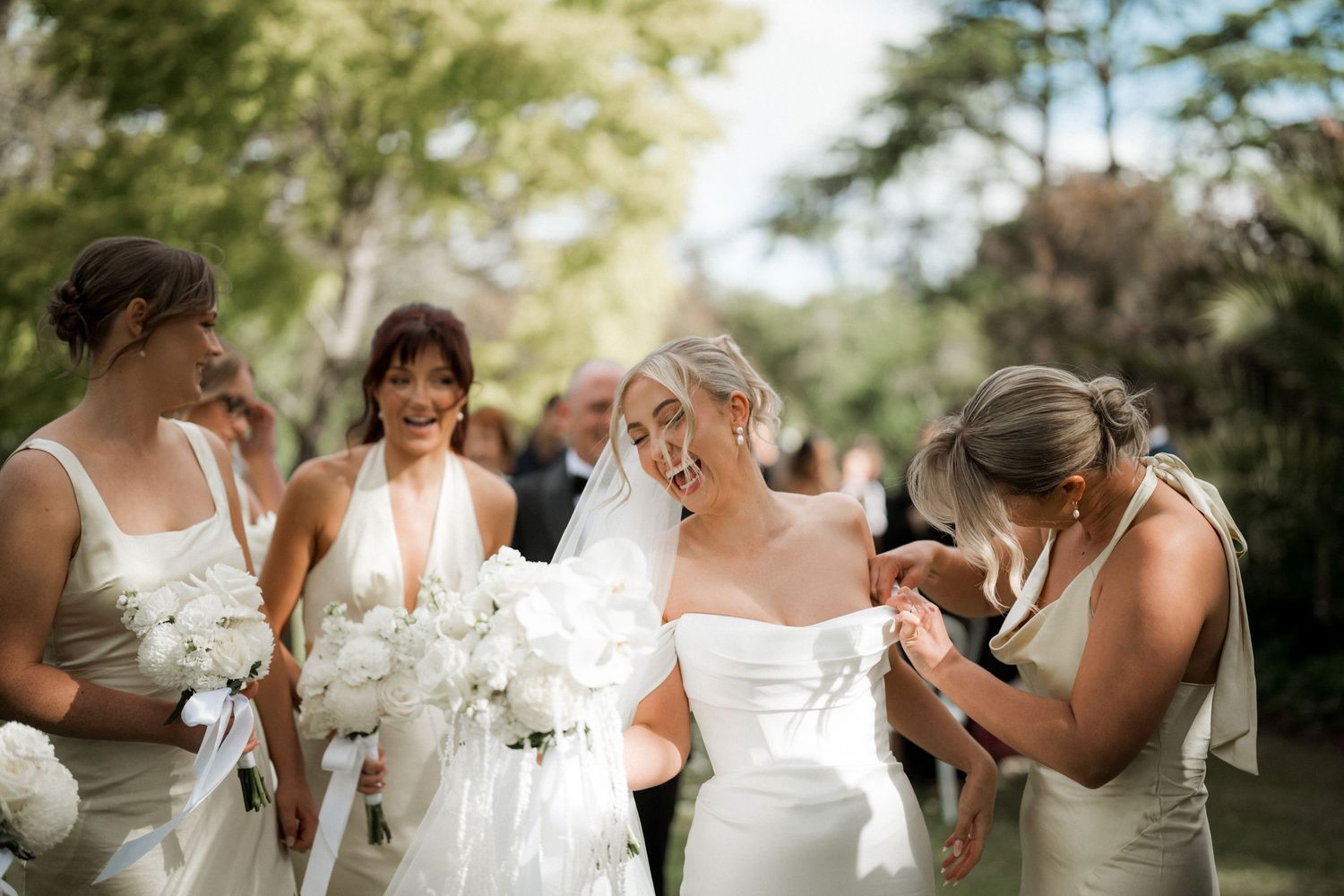 Wedding party wearing white dresses and holding bouquets pose together outdoors.