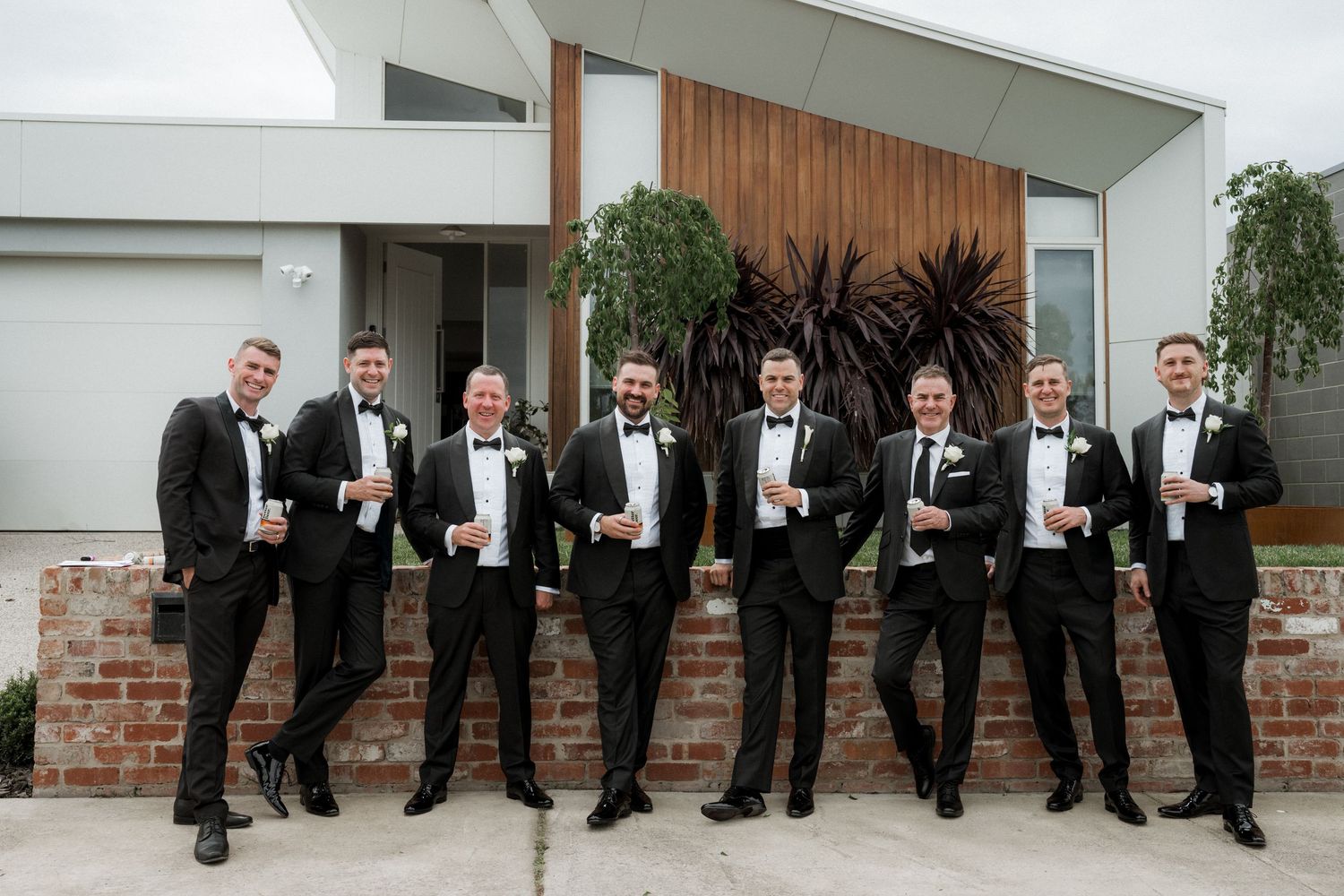 Groomsmen in black tuxedos pose playfully with their legs kicked up against a brick and white wall.