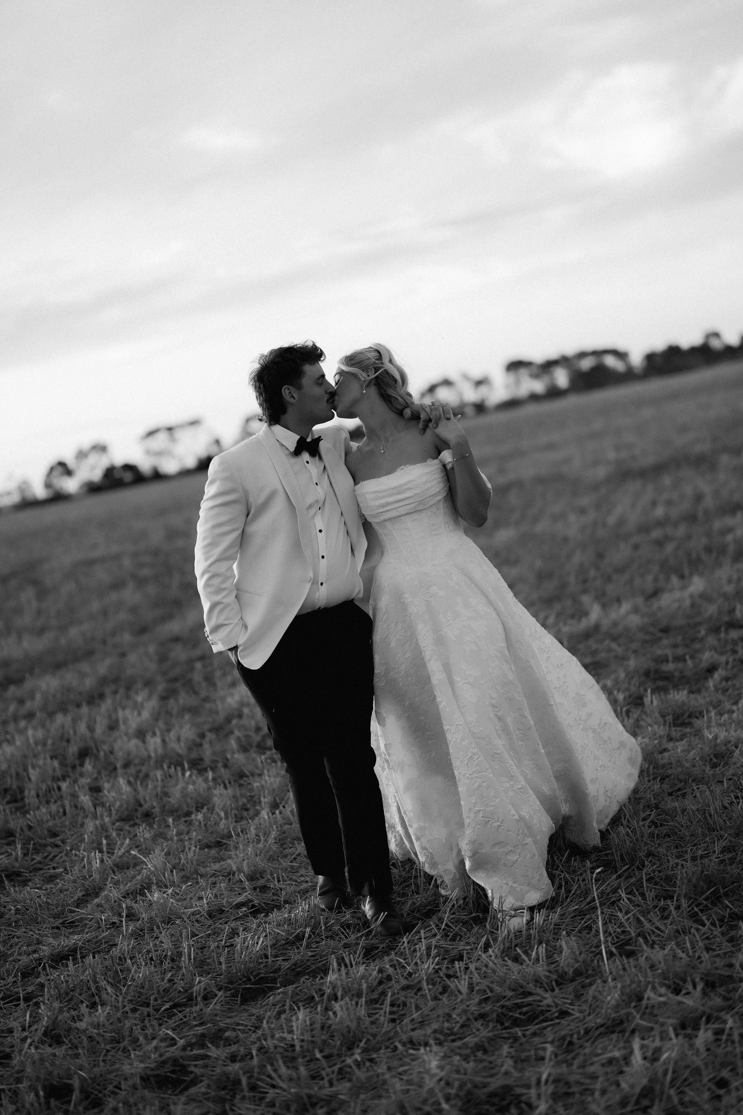 Bride and groom share a romantic kiss in an open field at dusk in a classic black and white wedding photo.