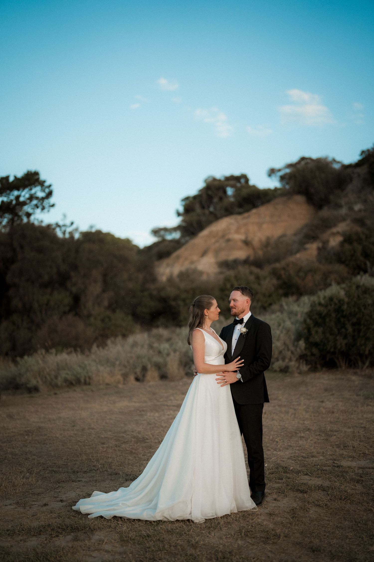 Newlyweds pose together on a hilltop against a bright blue sky.