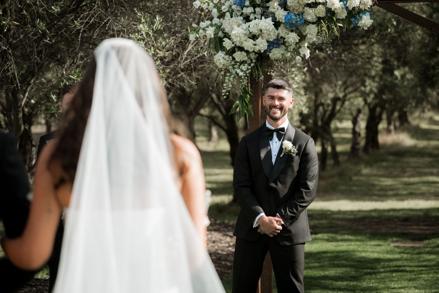 A bride and groom share a joyful moment under trees during their outdoor wedding ceremony.