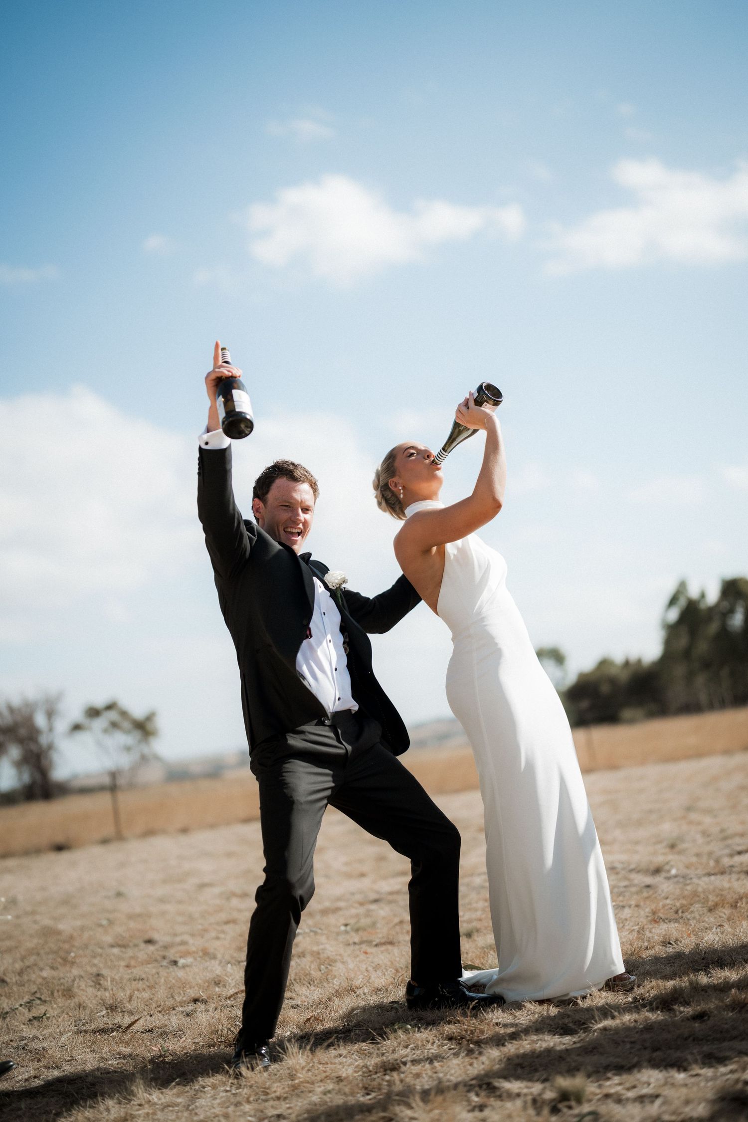 Playful wedding couple taking fun photos in an outdoor field against blue sky.