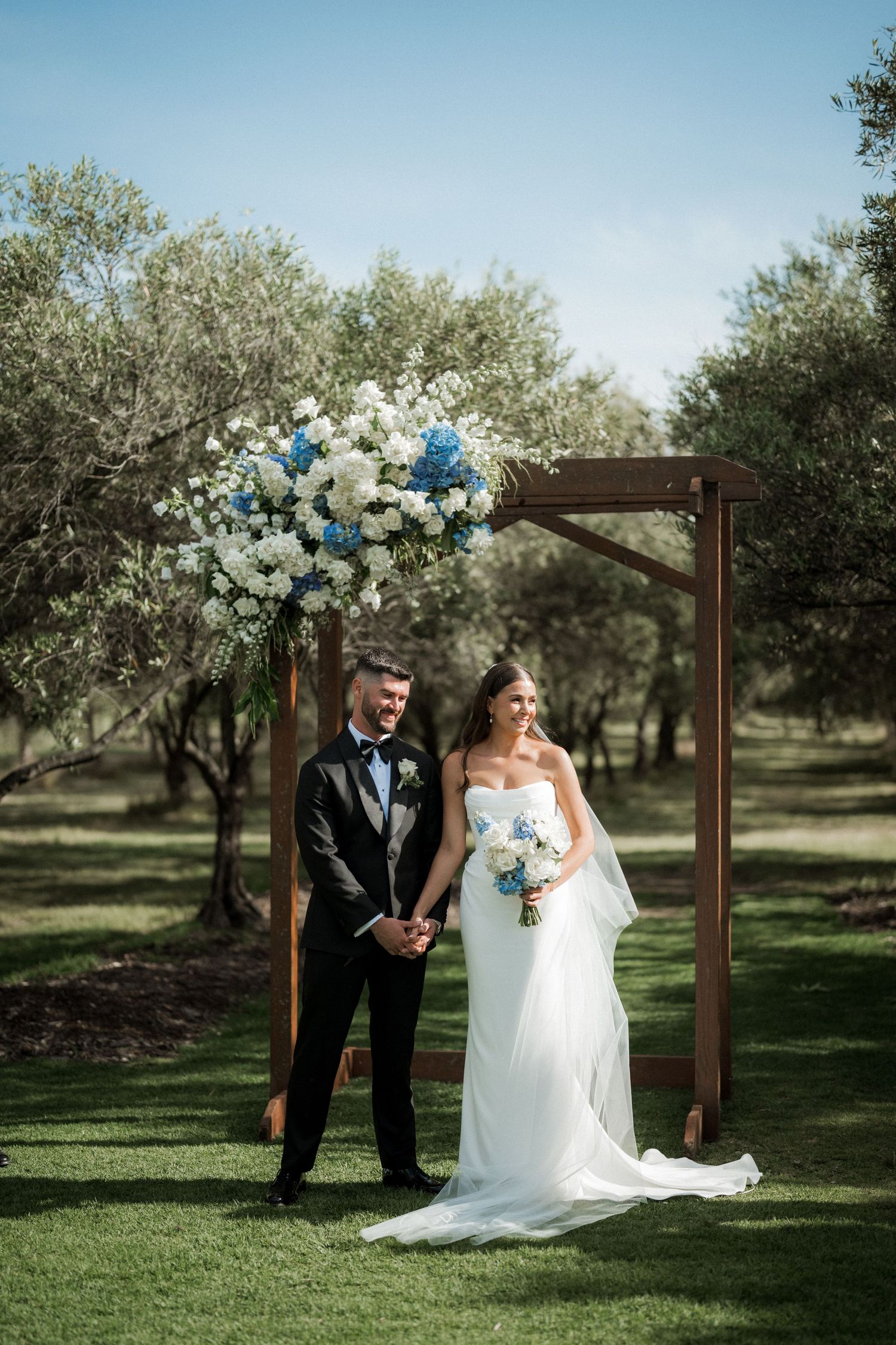An elegant wedding arch adorned with white and blue flowers stands in a serene olive grove setting.