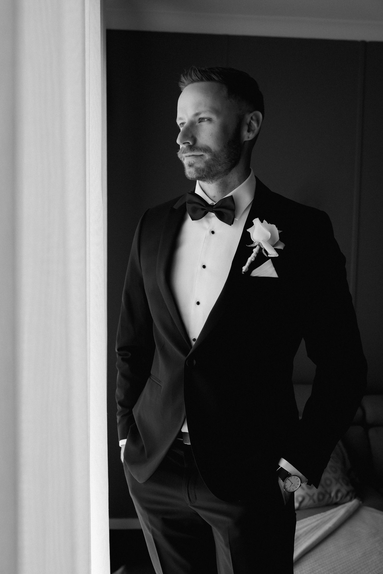 Elegant groom in classic black tuxedo and bow tie poses in dramatic lighting.