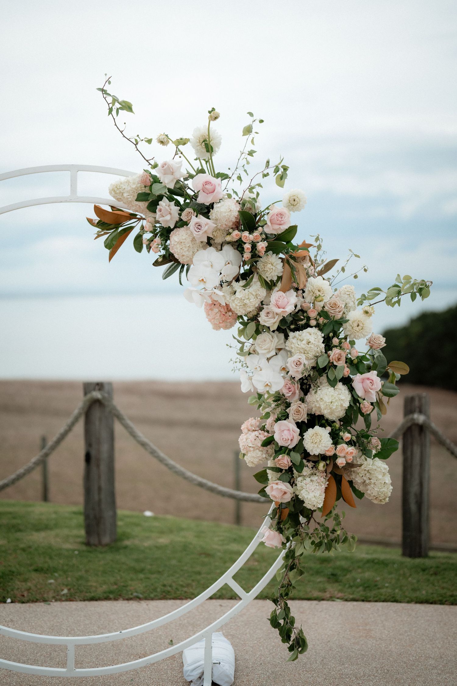 An elegant white and pink floral arch arrangement adorns a curved metal frame outdoors against a coastal backdrop.