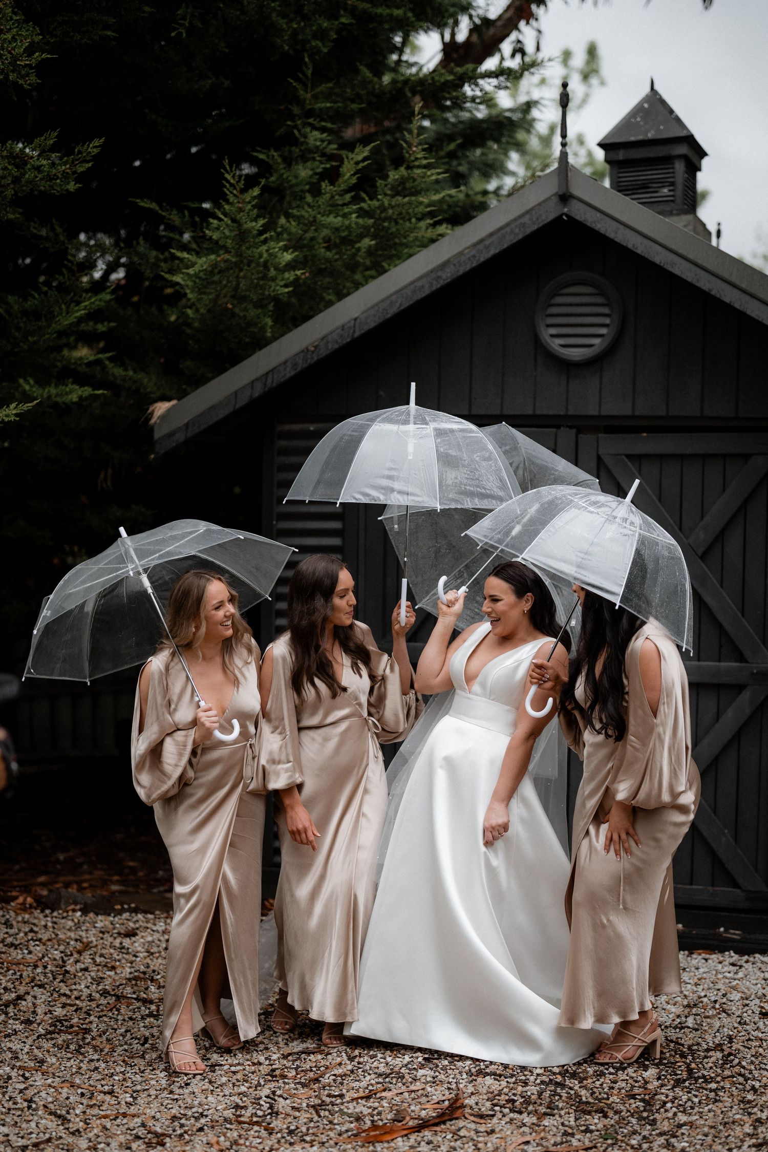 Group stands together holding clear umbrellas in formal wear against rustic barn on overcast wedding day.
