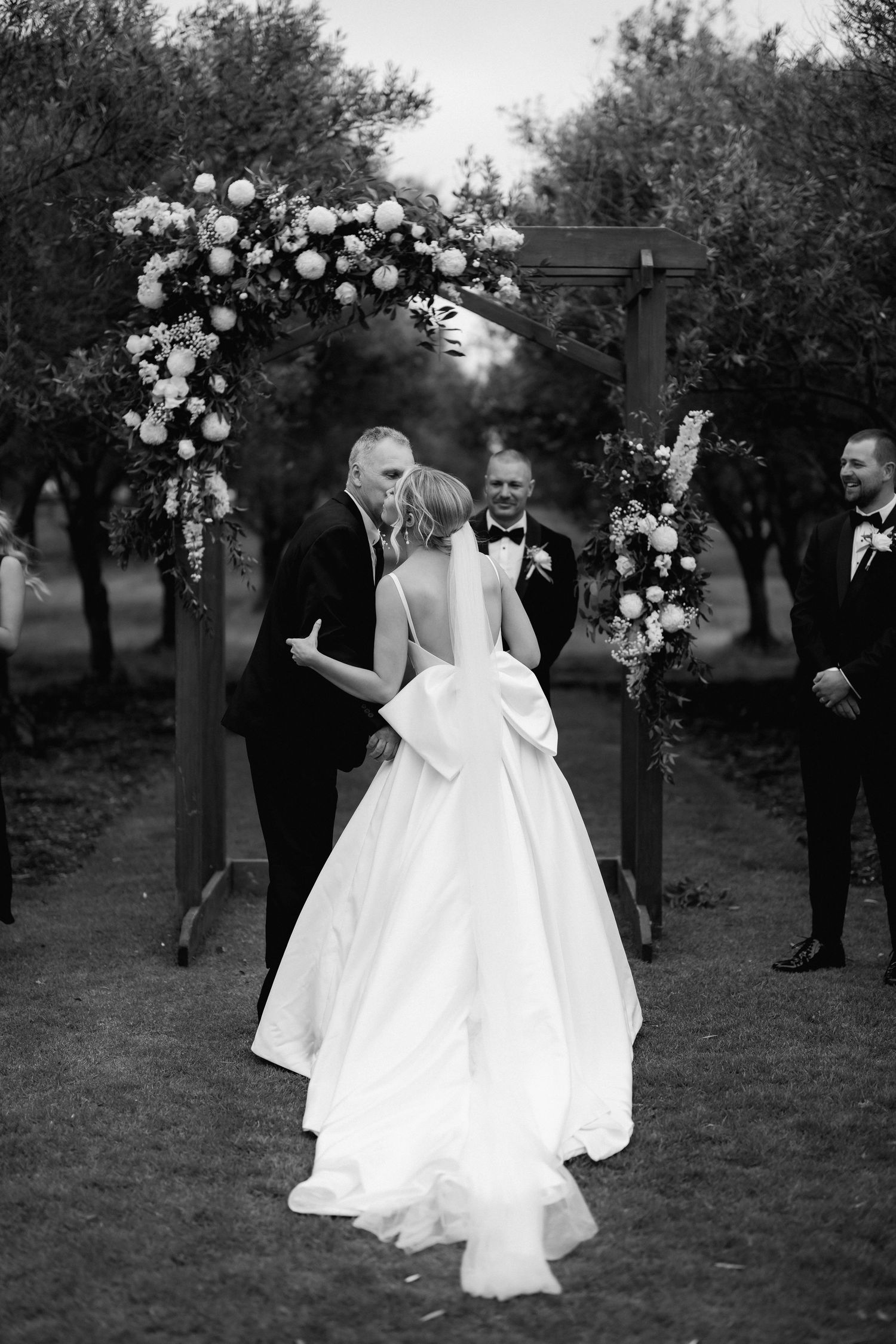 Wedding ceremony under a floral arch with people gathered in a garden setting.