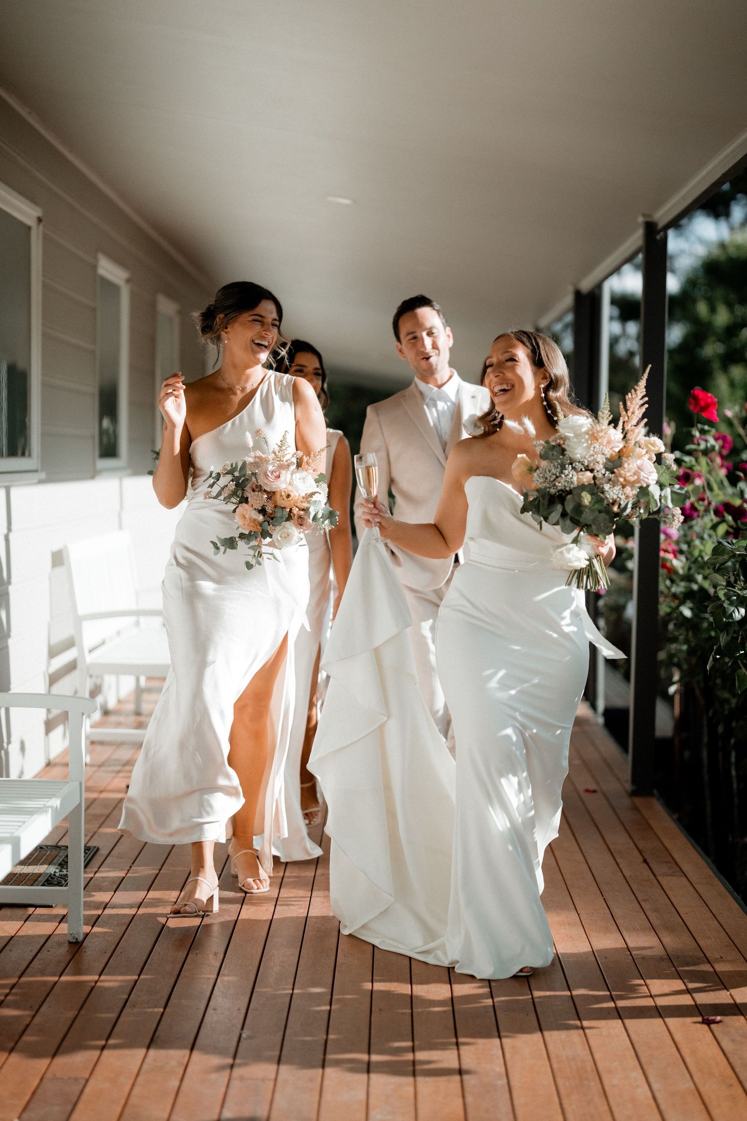 Group in white dresses pose playfully on a wooden porch during a special occasion.