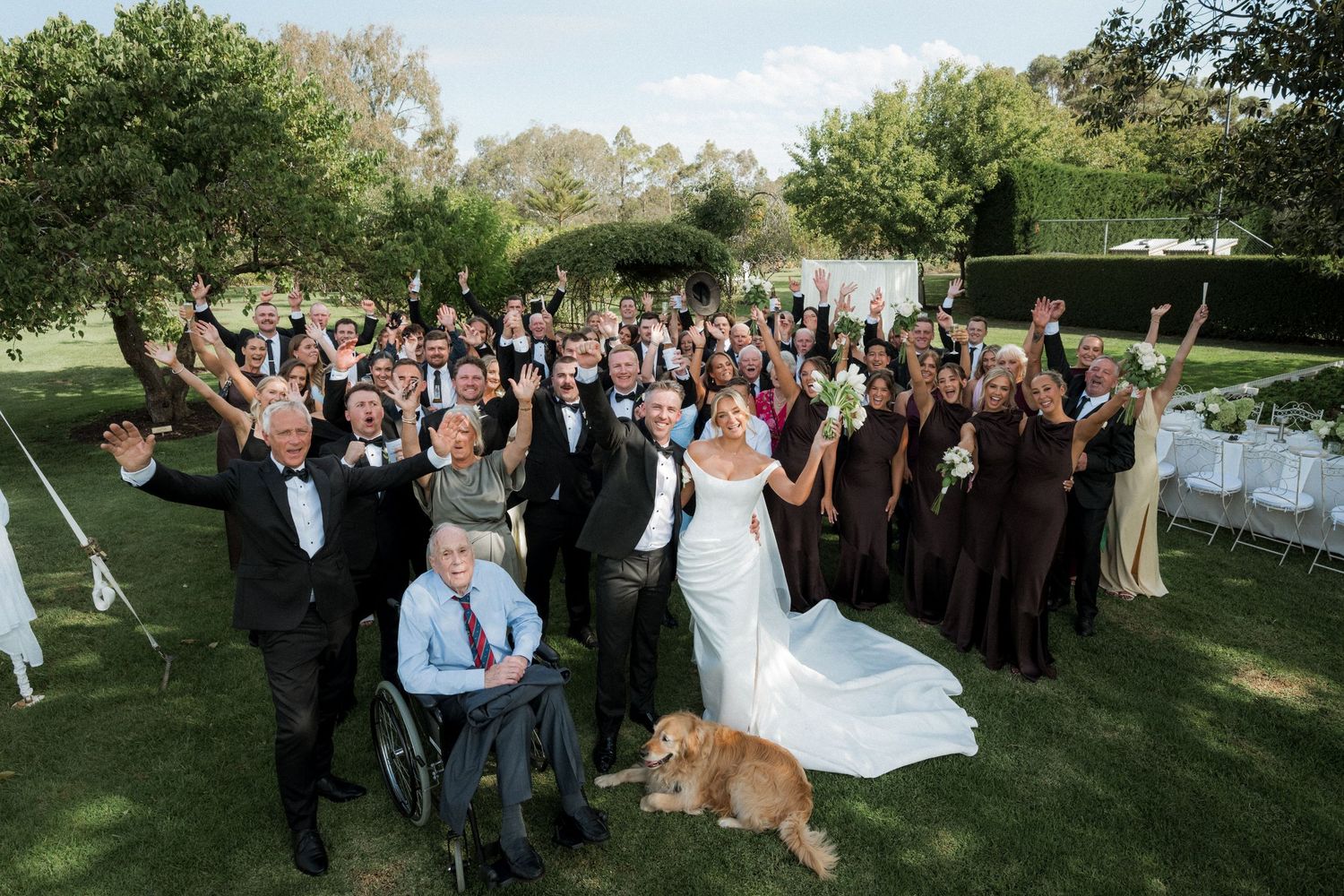 Large wedding party poses together on green lawn with dog during outdoor garden ceremony.