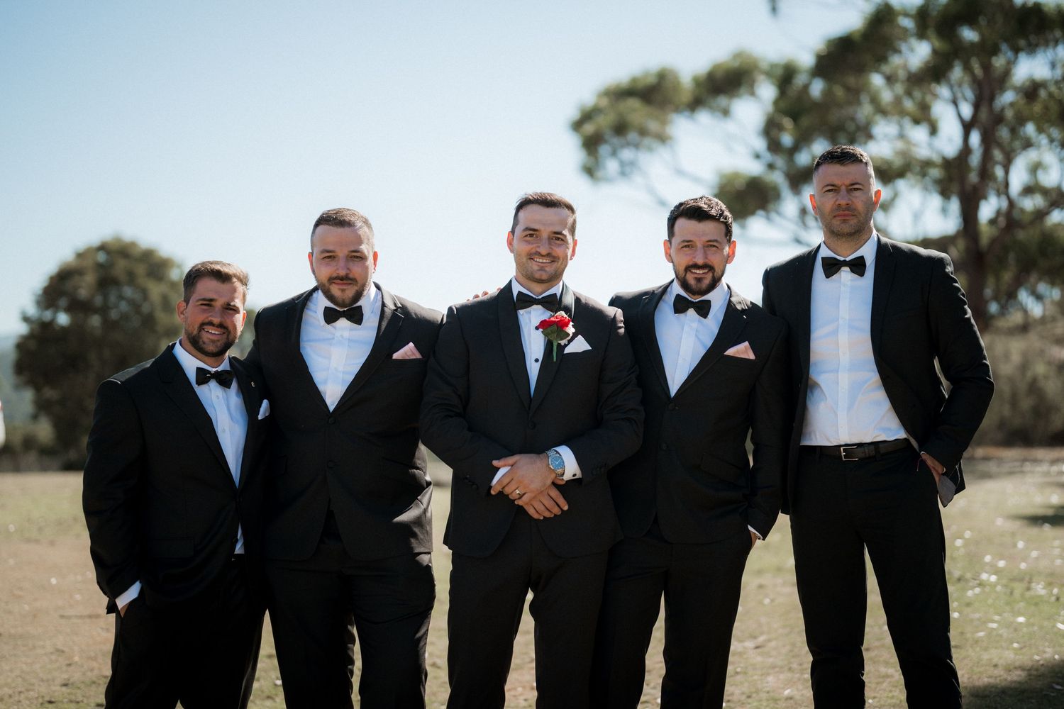 Groomsmen in black tuxedos and bowties pose together outdoors for a wedding photo.