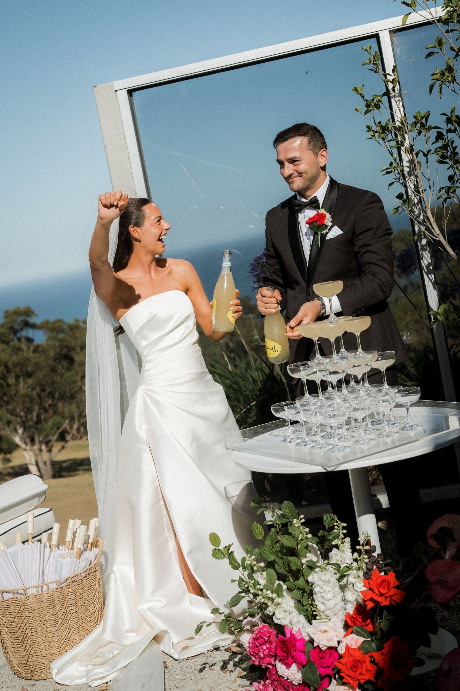 Newlyweds celebrate by champagne tower at outdoor reception with white and red floral decorations.