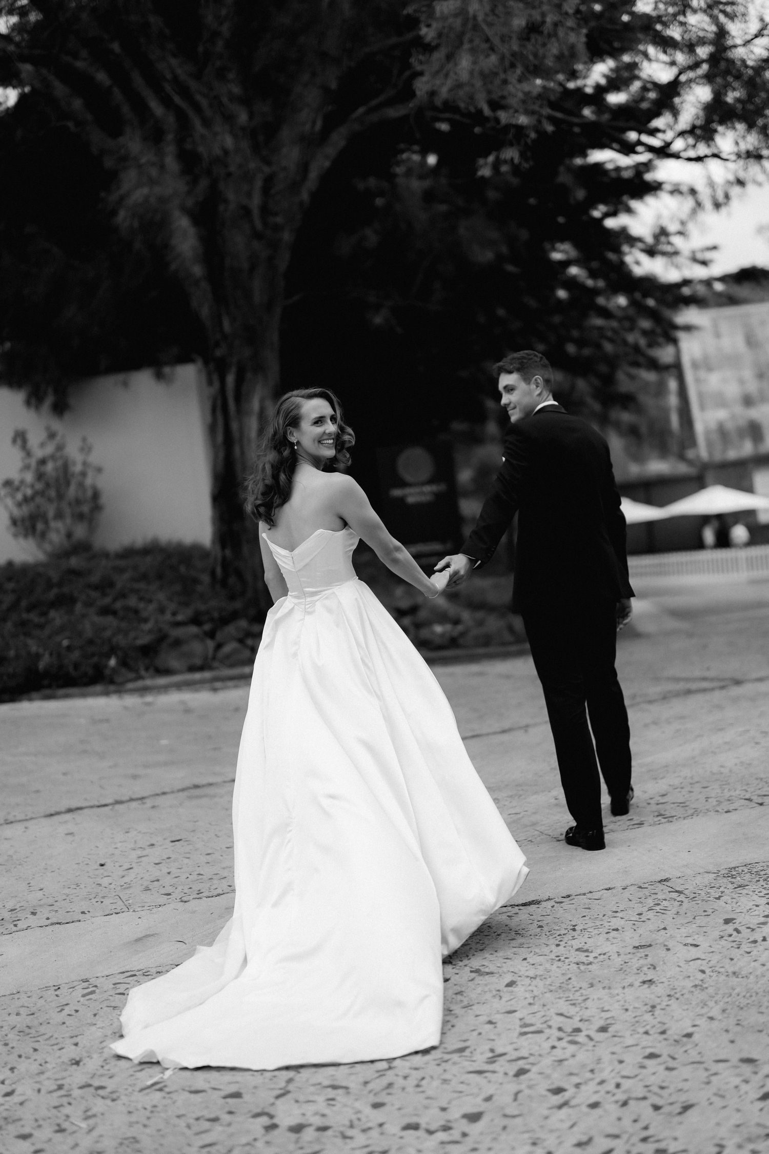 Black and white photo of a couple walking together on a path, with the bride's white dress flowing behind.