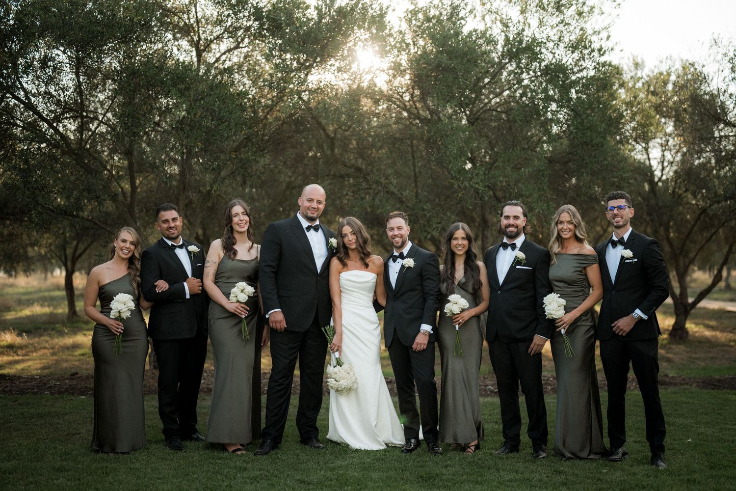 Wedding party standing in line outdoors with trees in background during golden hour.