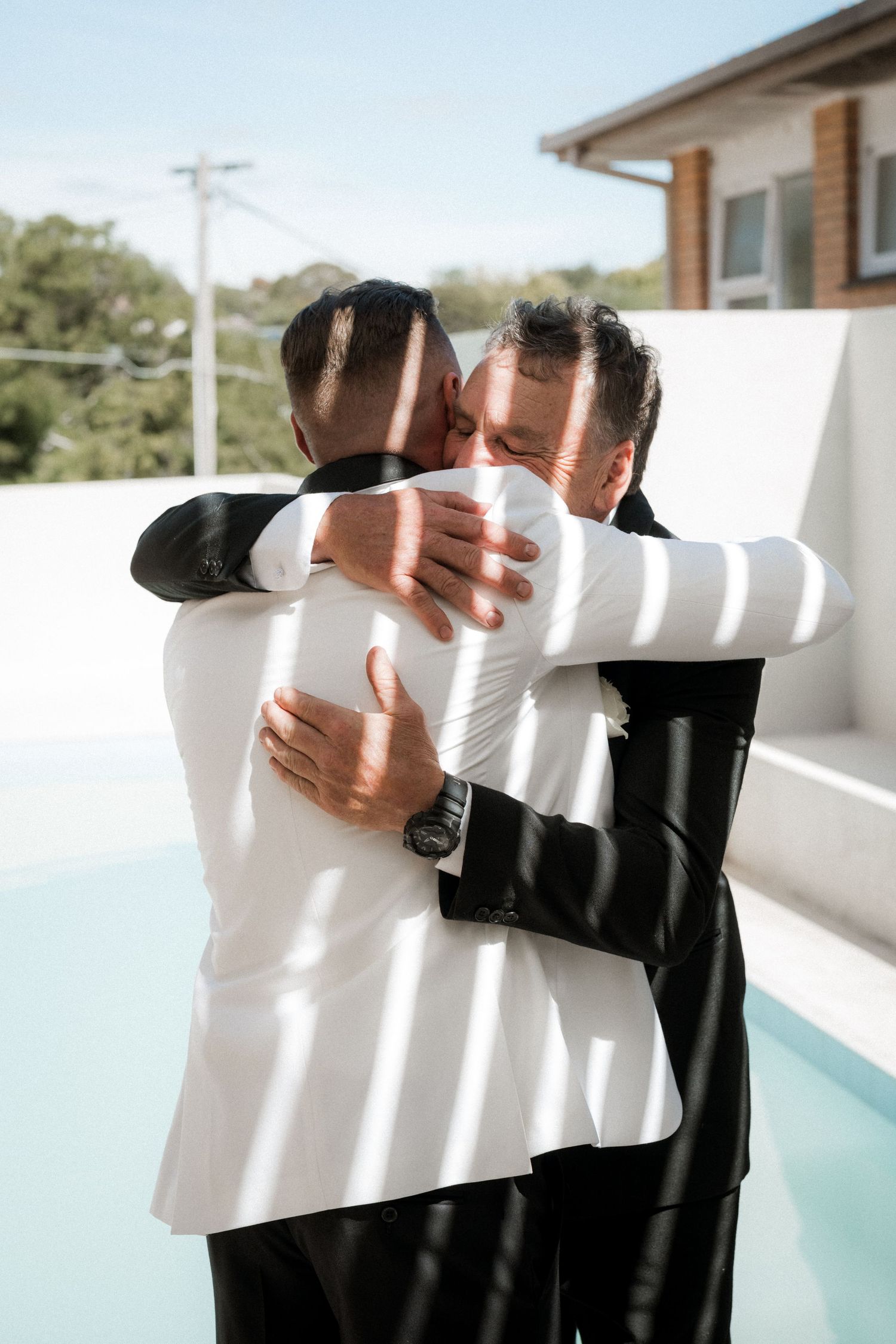 Two people in formal attire share an emotional embrace by a swimming pool on a sunny day.