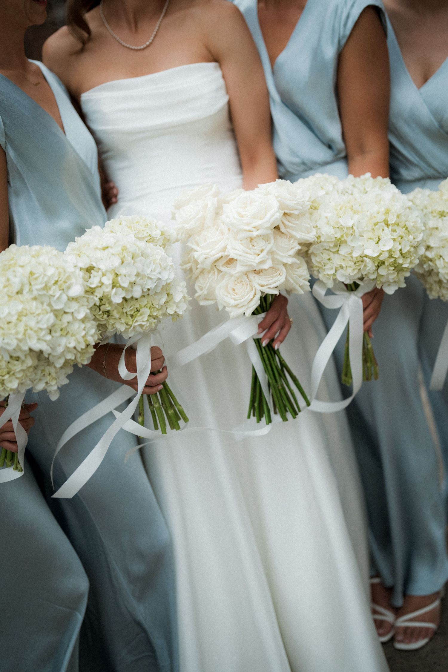 A row of bridesmaids in light blue dresses holding elegant white hydrangea bouquets with satin ribbons.