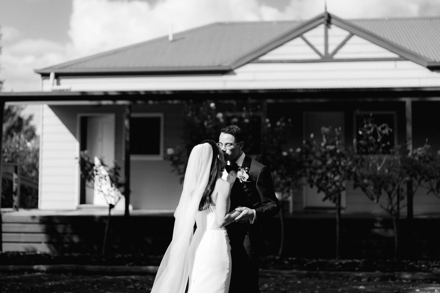 Black and white wedding photo of bride and groom sharing a tender moment outside a classic country homestead.