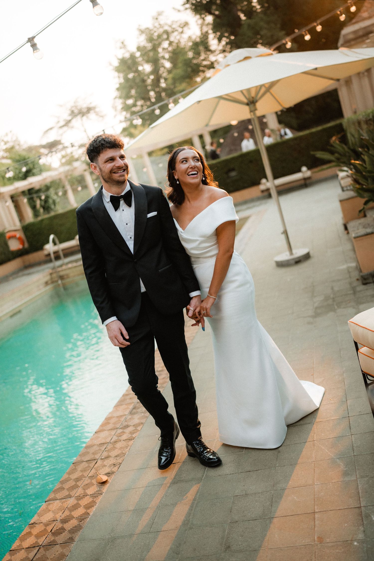 A newlywed couple laughs together by a pool at sunset during their wedding reception.