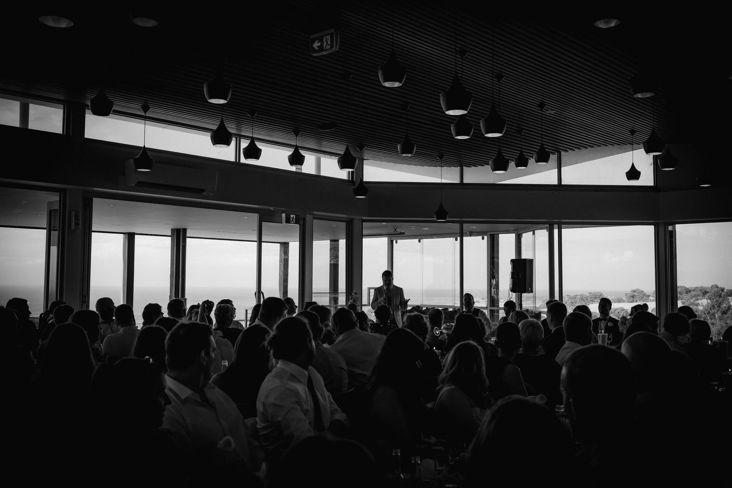 Silhouetted wedding guests gather in a modern venue with floor-to-ceiling windows and dramatic lighting.