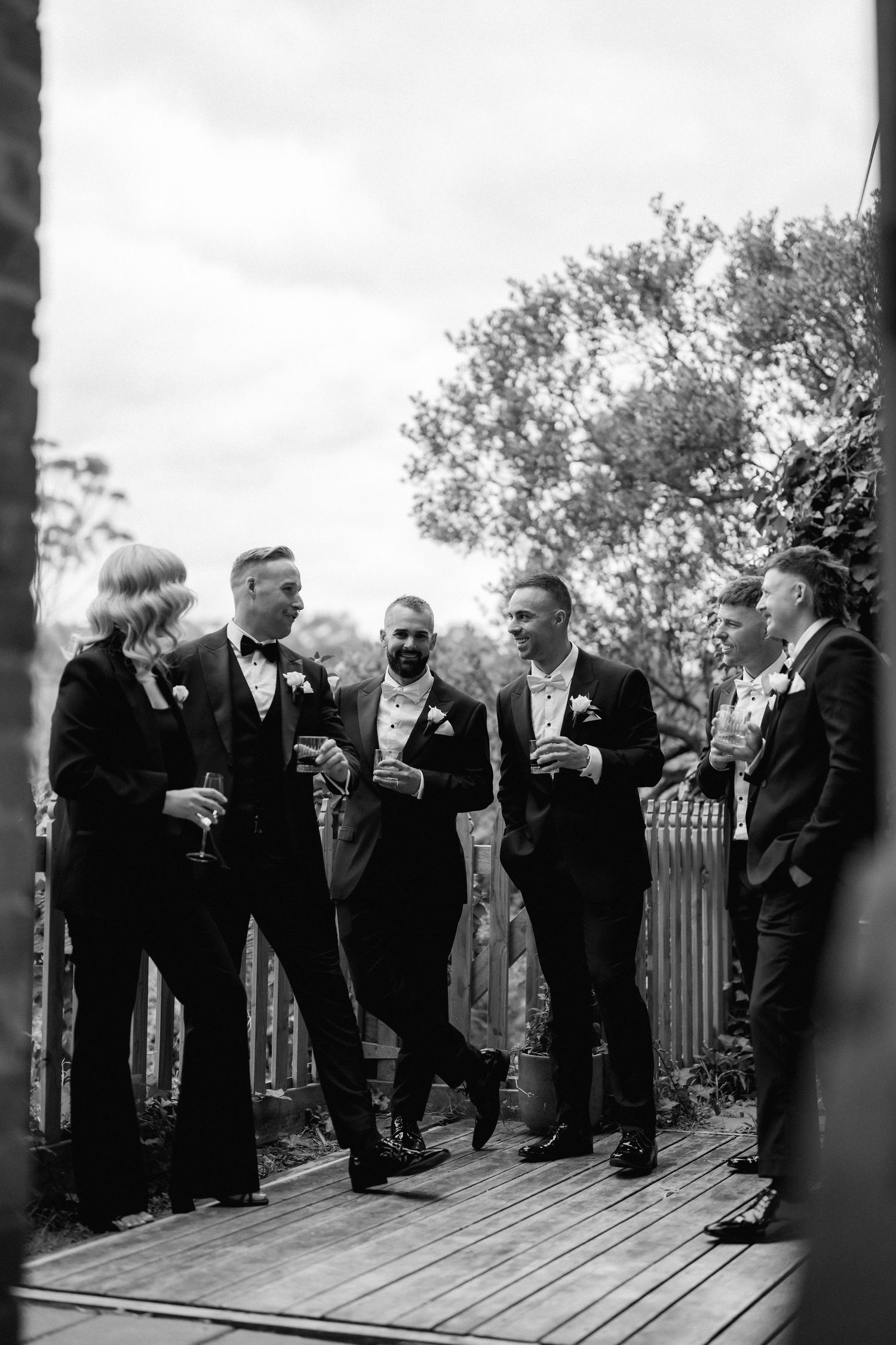 Groomsmen in black tuxedos sharing drinks and laughing together on a wooden deck in black and white photography.