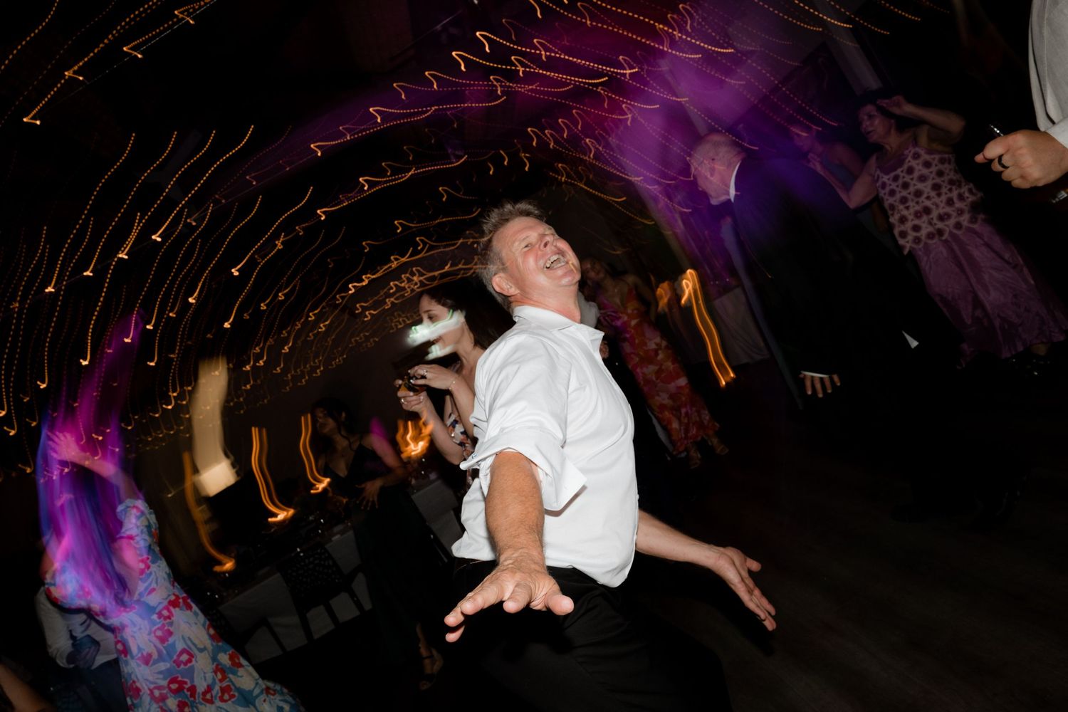 A wedding guest dances energetically under purple mood lighting in a darkened reception venue.