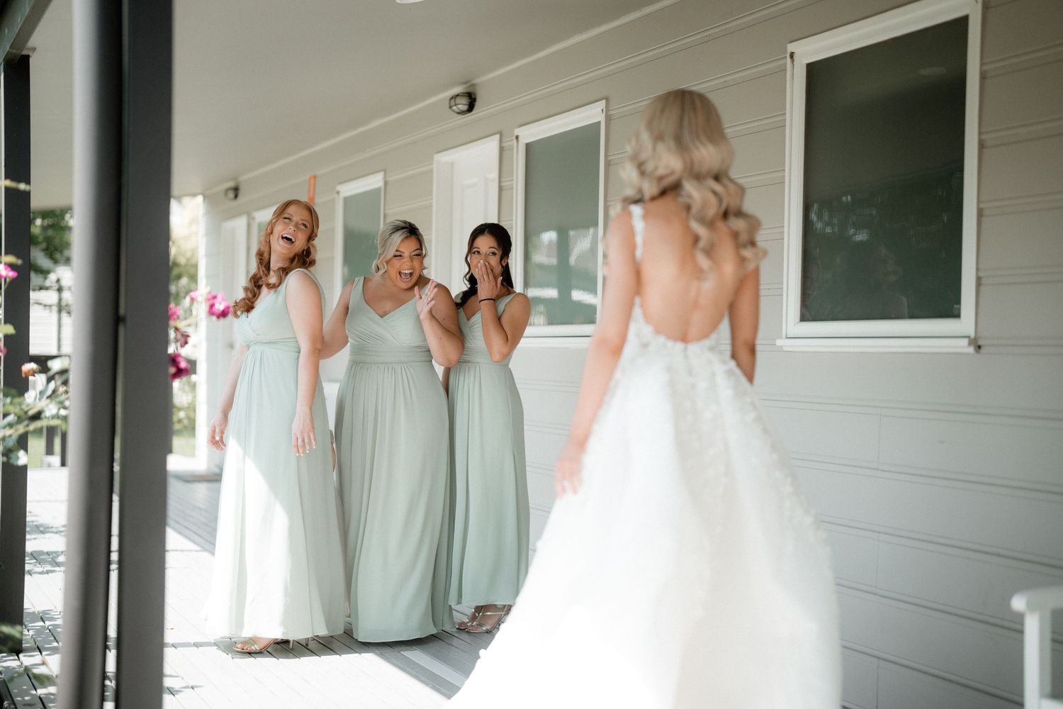 Bridesmaids in light sage green dresses react with joy seeing bride in white backless wedding gown on porch.
