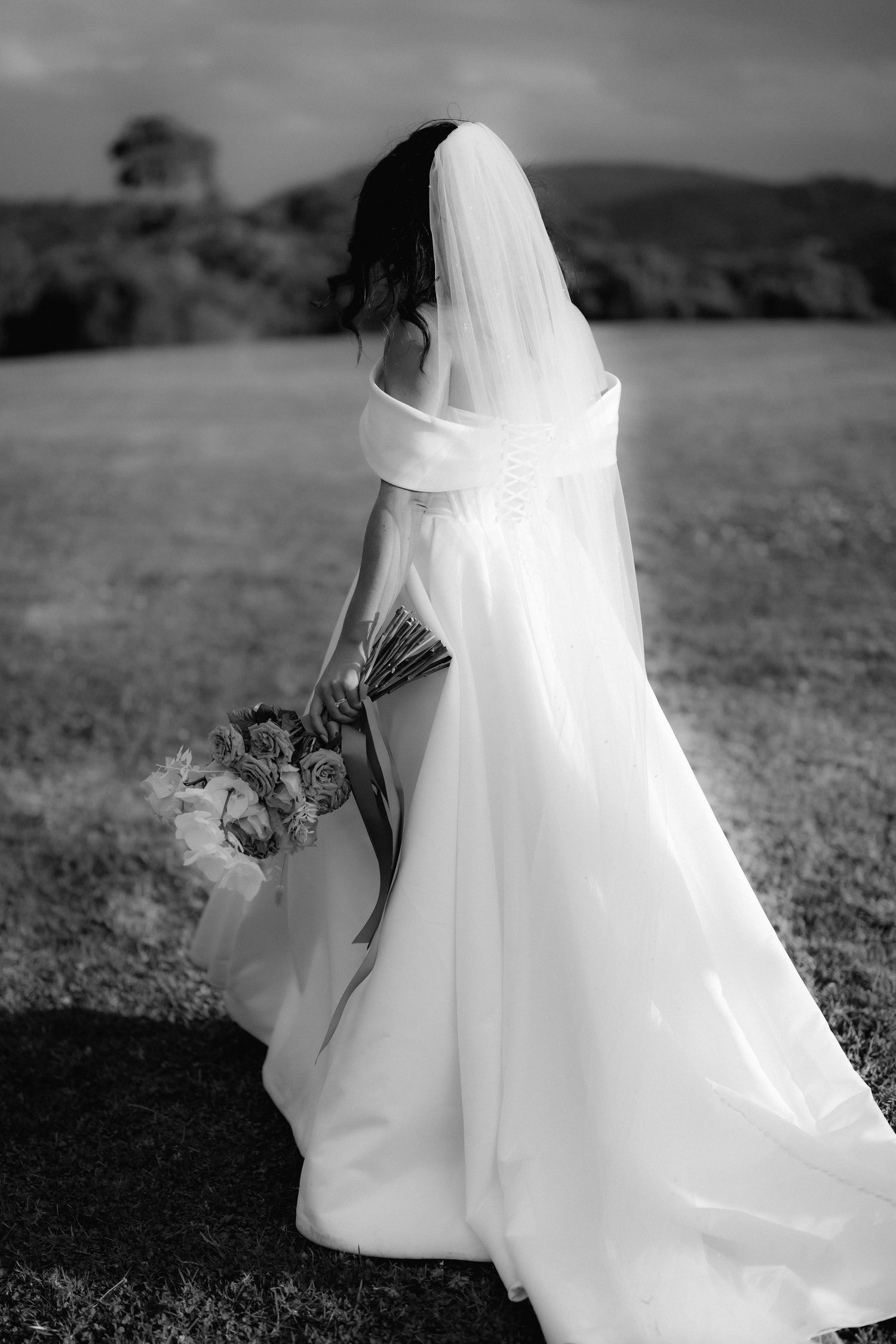 Black and white photograph of a bride in a flowing white wedding dress holding a bouquet on a hilltop landscape.