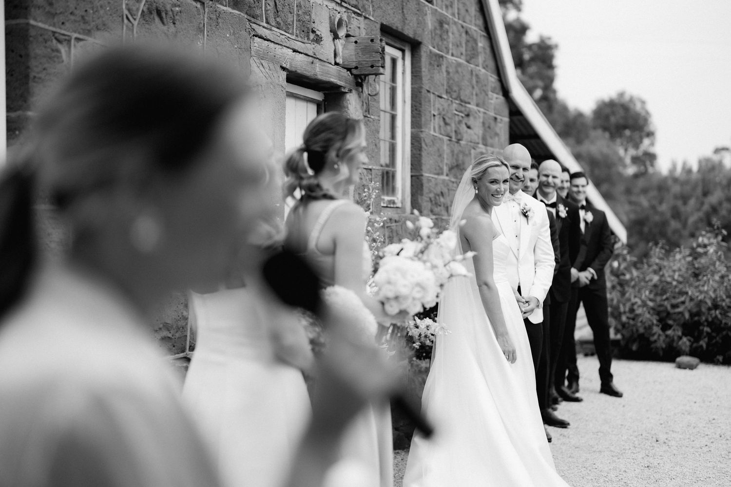 A vintage black and white photograph showing a wedding party gathering outside a rustic stone building.