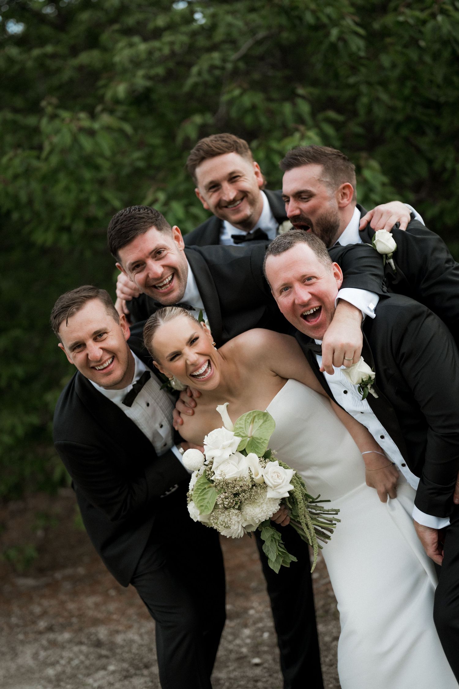 Wedding party members in black tuxedos and white dress share candid laughing moments during outdoor photo session.