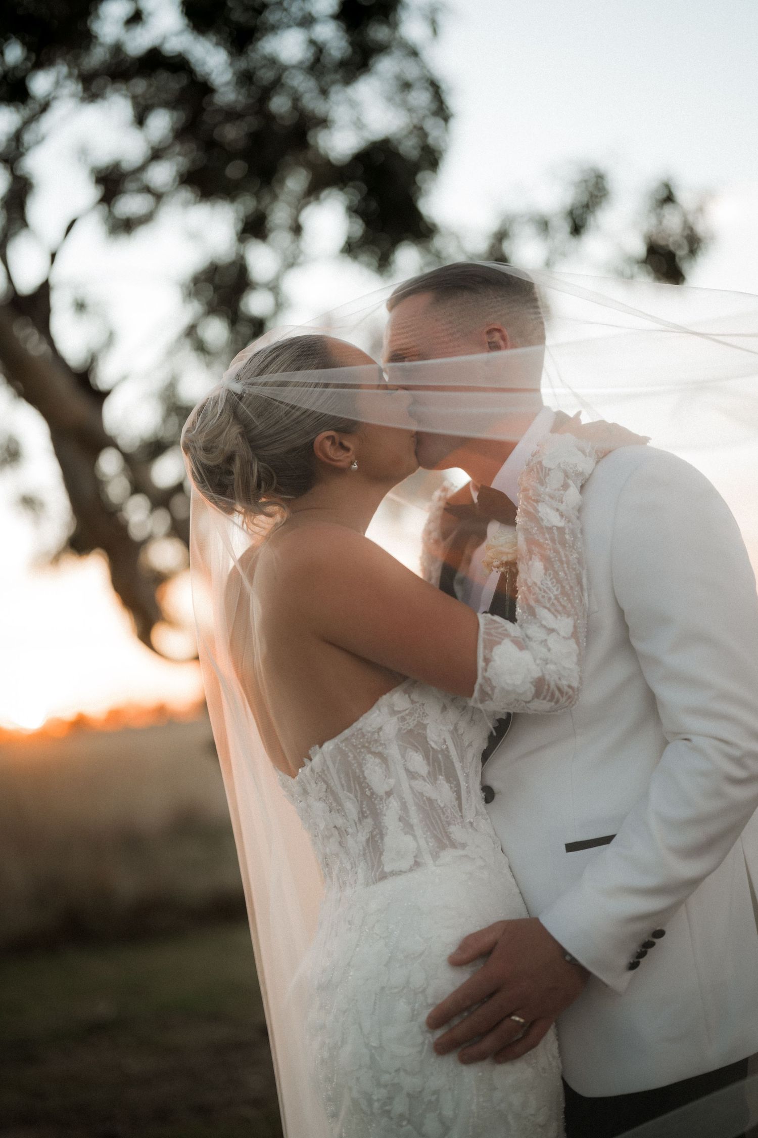 Romantic wedding couple sharing an intimate moment under a veil during sunset with trees in background.