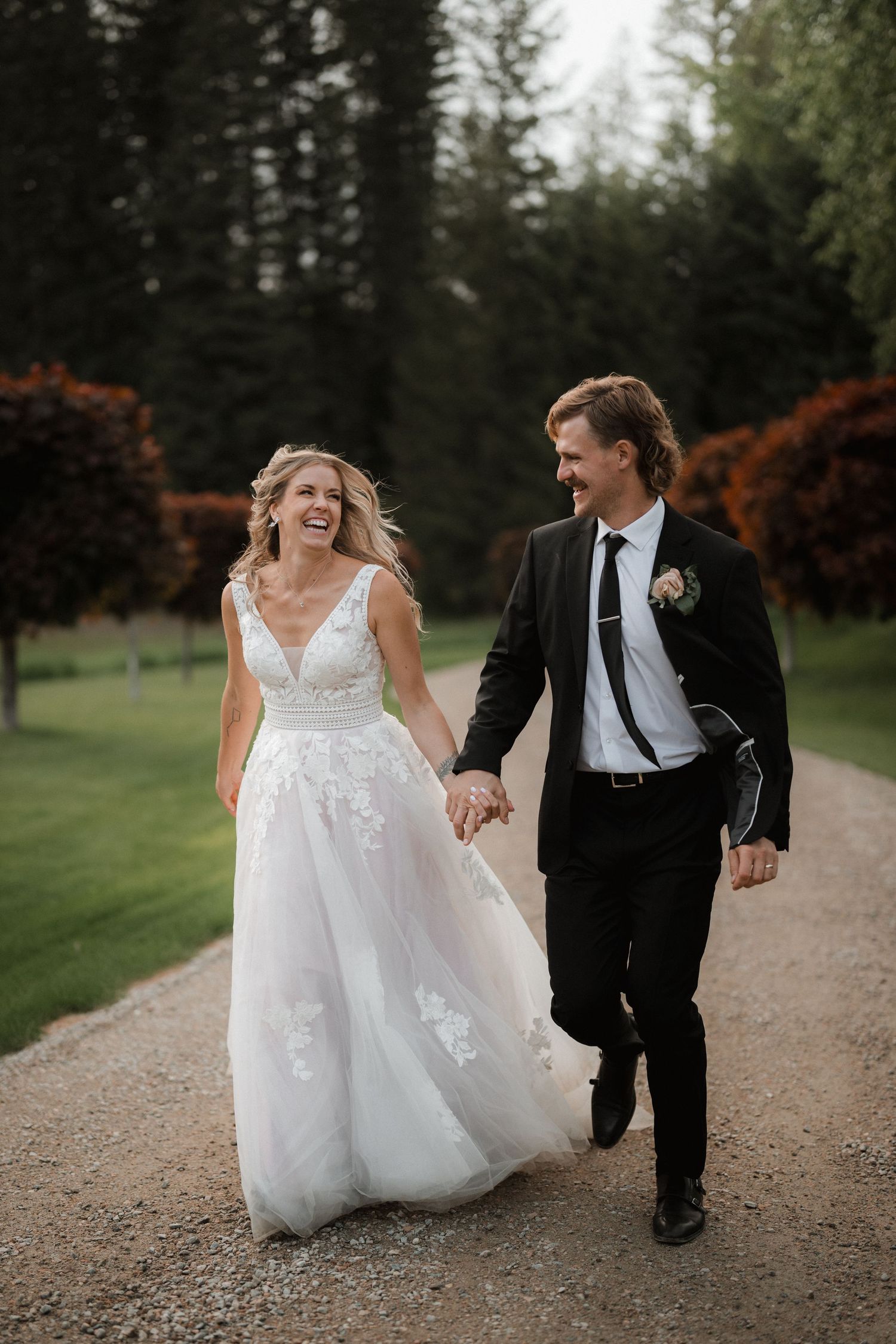 A joyful couple in wedding attire runs and laughs together on a gravel path surrounded by evergreen trees.