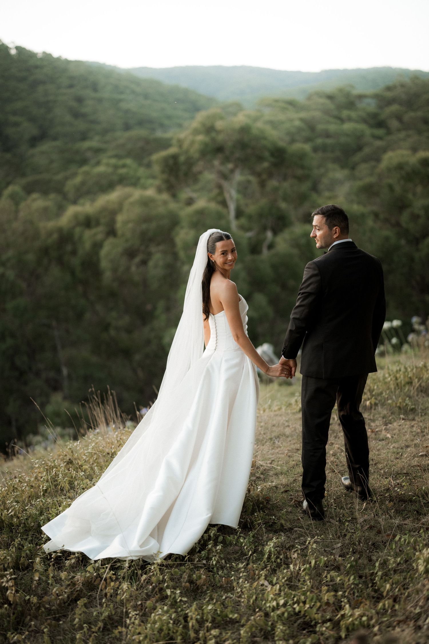 A wedding couple poses together on a hillside surrounded by lush green mountains and vegetation.