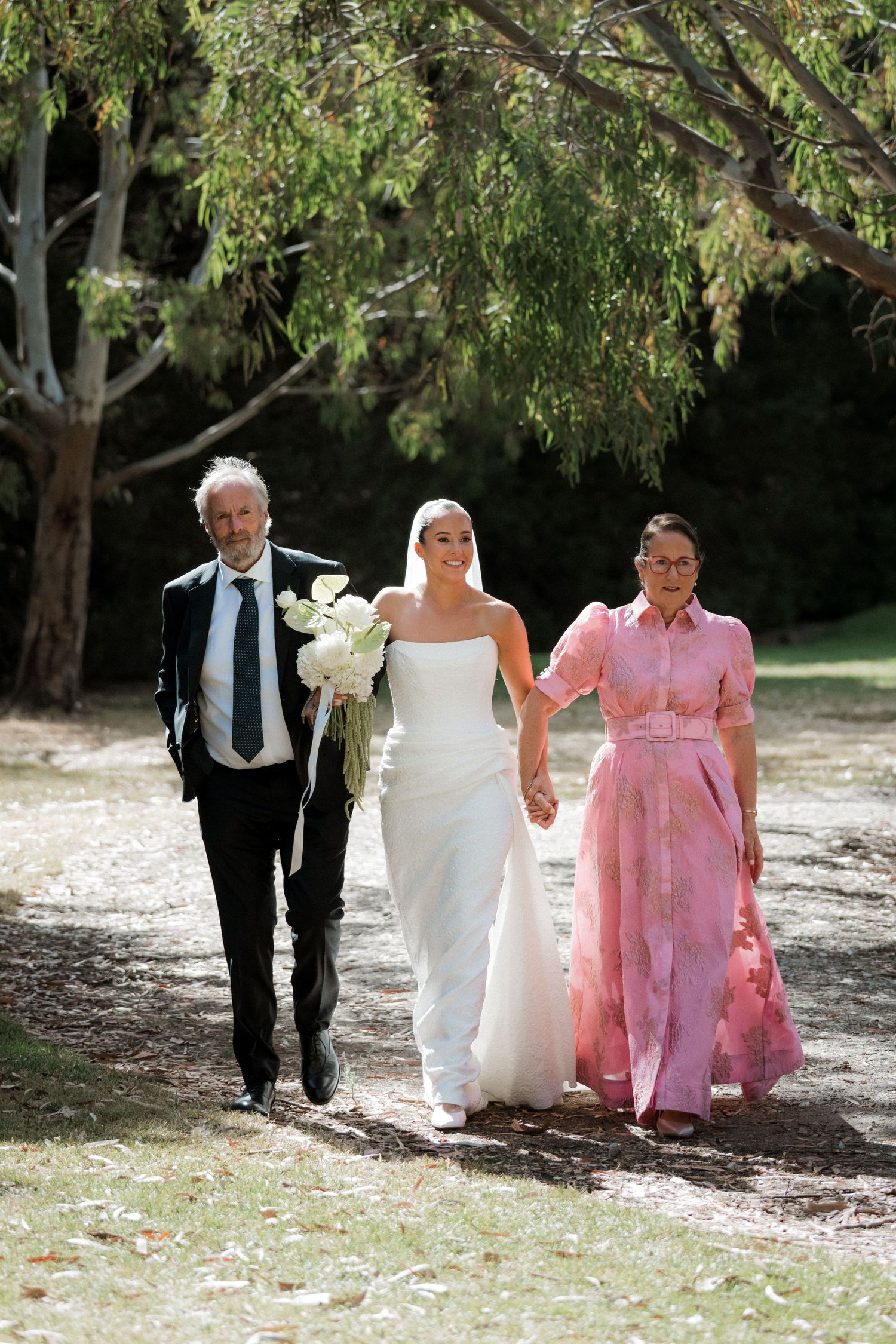 Bride in white gown holding bouquet walks with parents through sunlit garden path on her wedding day.
